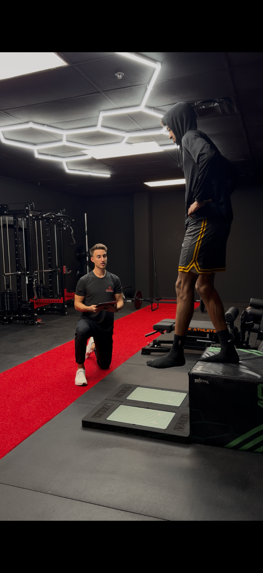 Personal trainer kneeling with clipboard talking to a young man on a box in a gym.