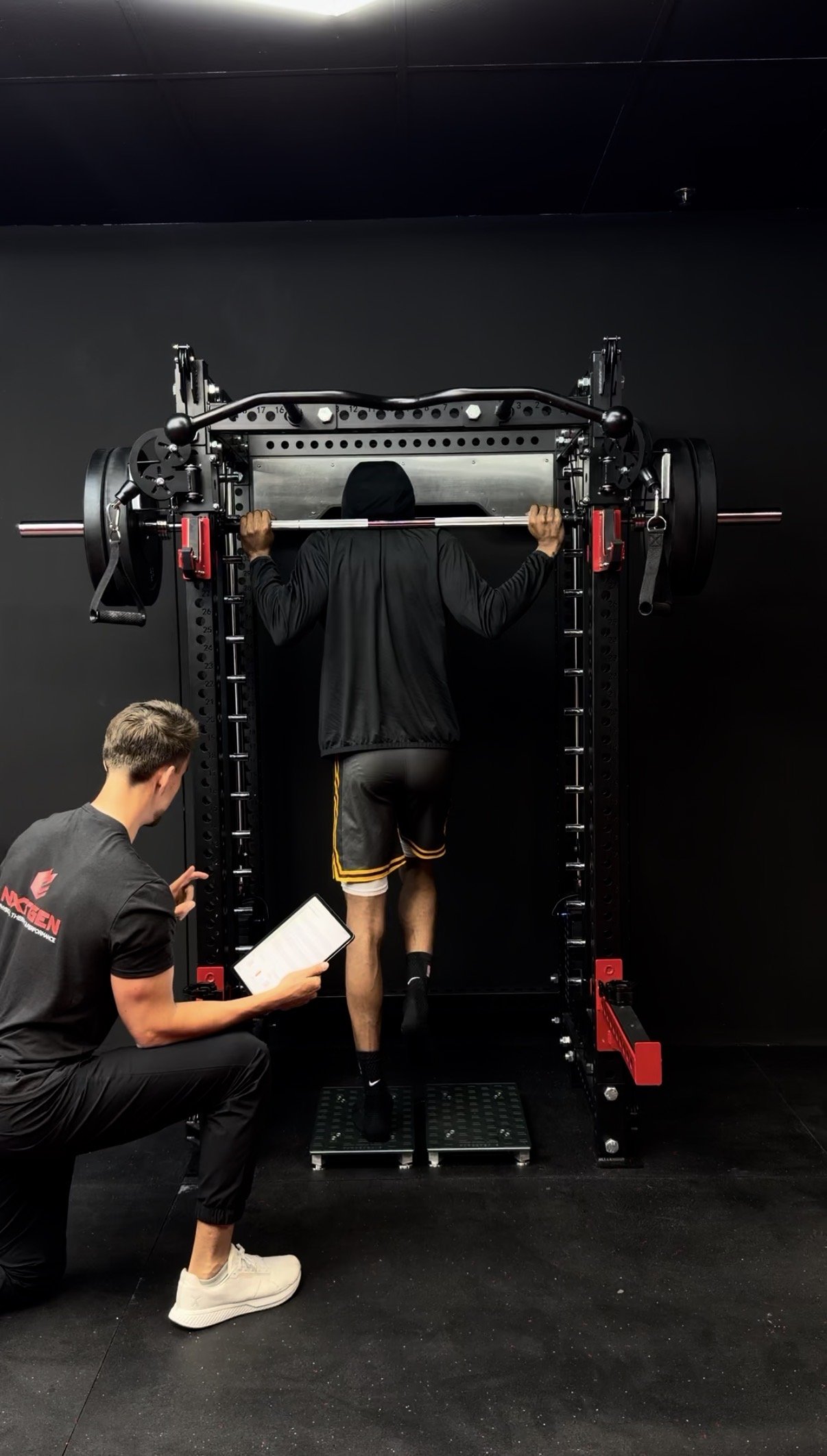 A man wearing workout clothes performs a strength test on a Smith machine while a trainer observes and takes notes, in a gym with black walls.