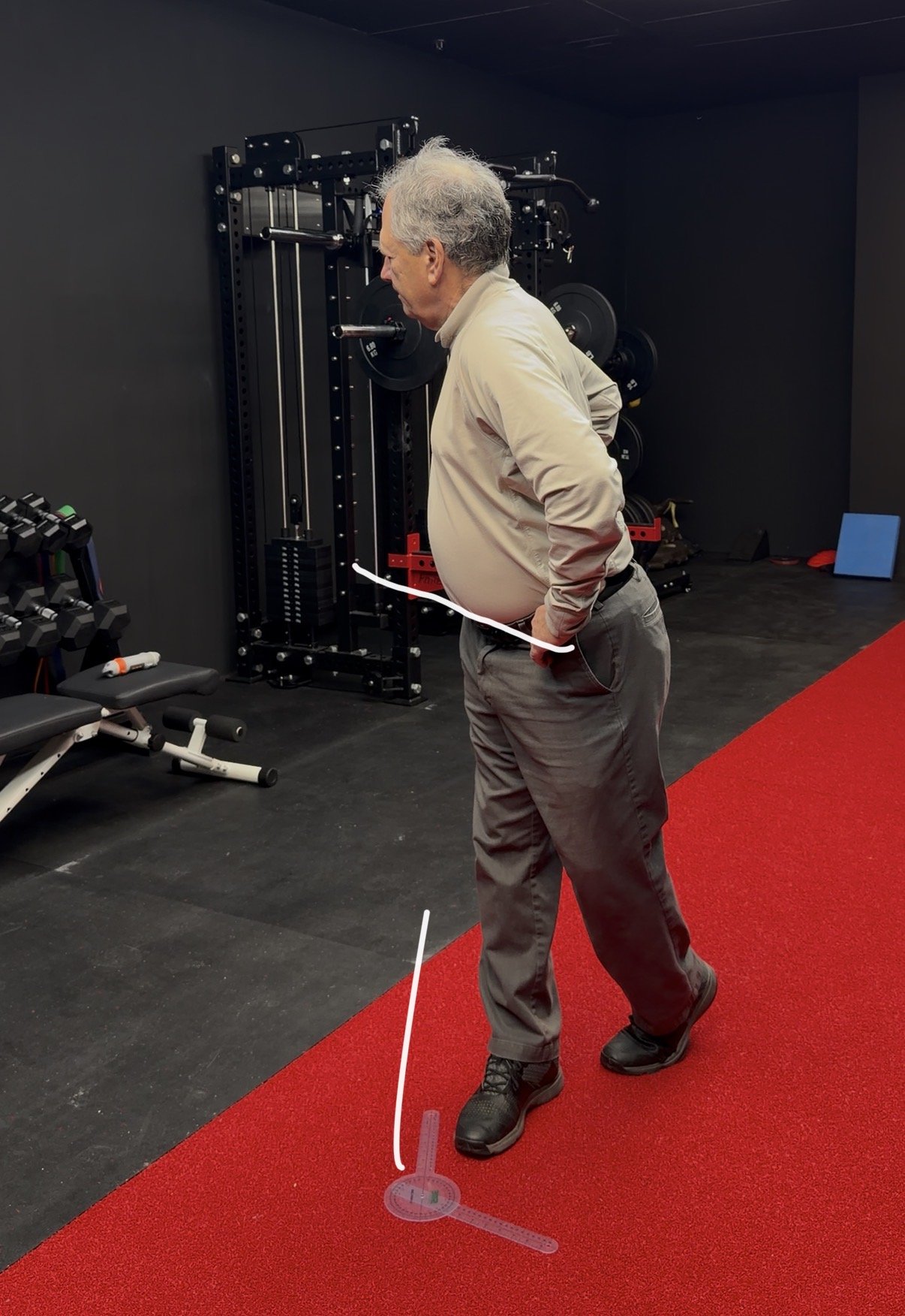 A man with gray hair, wearing a beige jacket, gray pants, and black shoes, walking on a red carpet in a gym. There are fitness equipment and black walls in the background.