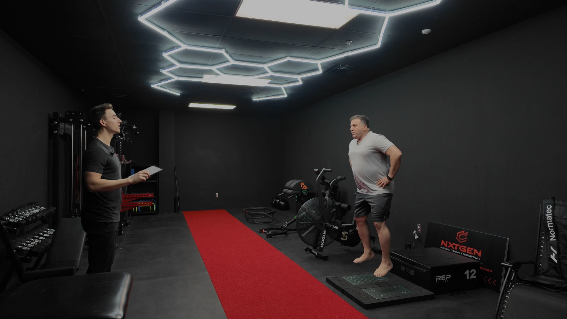 A man is doing a fitness assessment on a sensor platform in a gym with black walls and modern ceiling lights, while a trainer or coach stands nearby holding a clipboard and watching him.