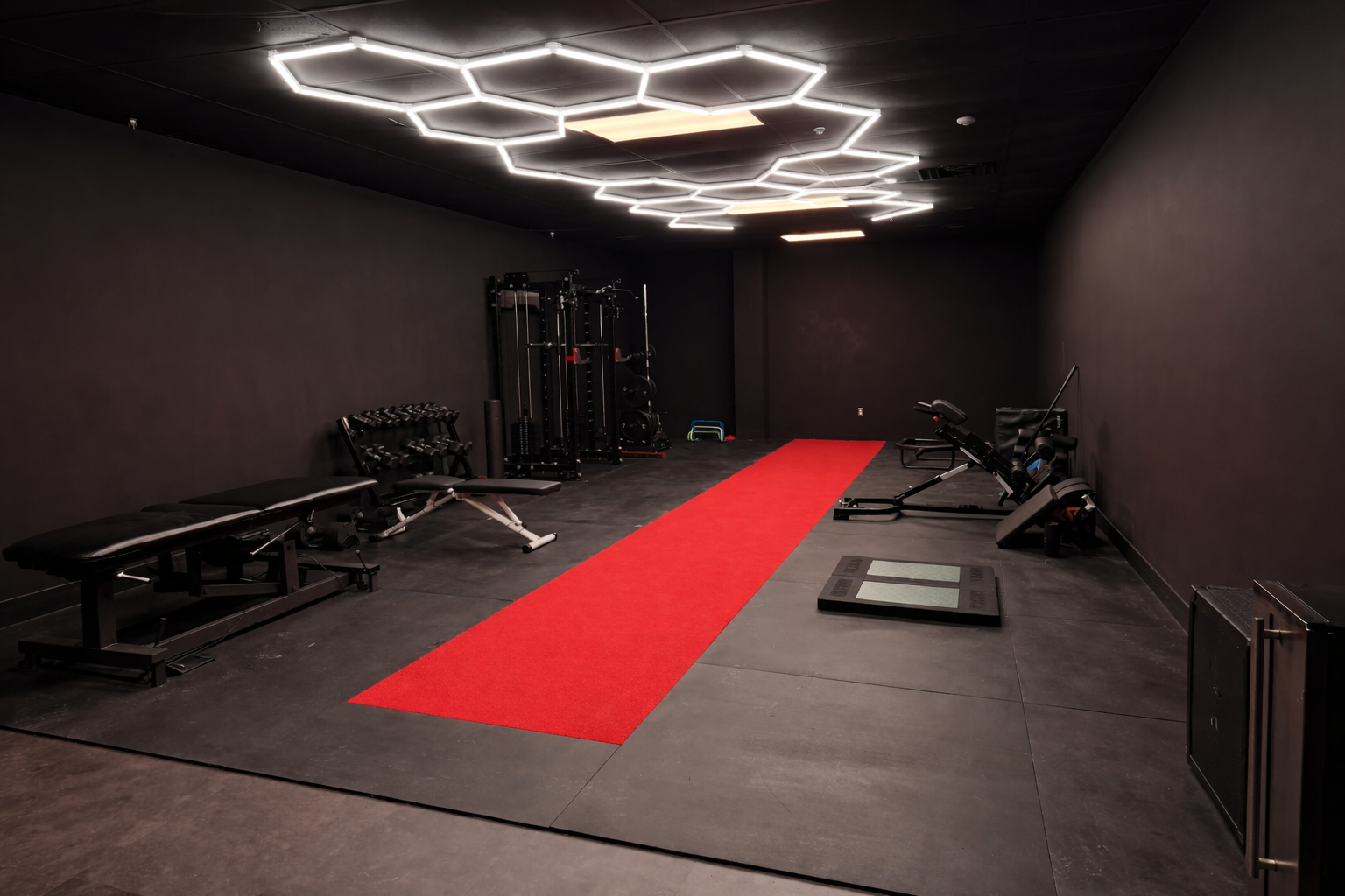 Empty gym room with black walls, a red running track in the center, and various fitness equipment including a bench, dumbbells, weight racks, and a stair climber. Modern hexagonal LED ceiling lights.