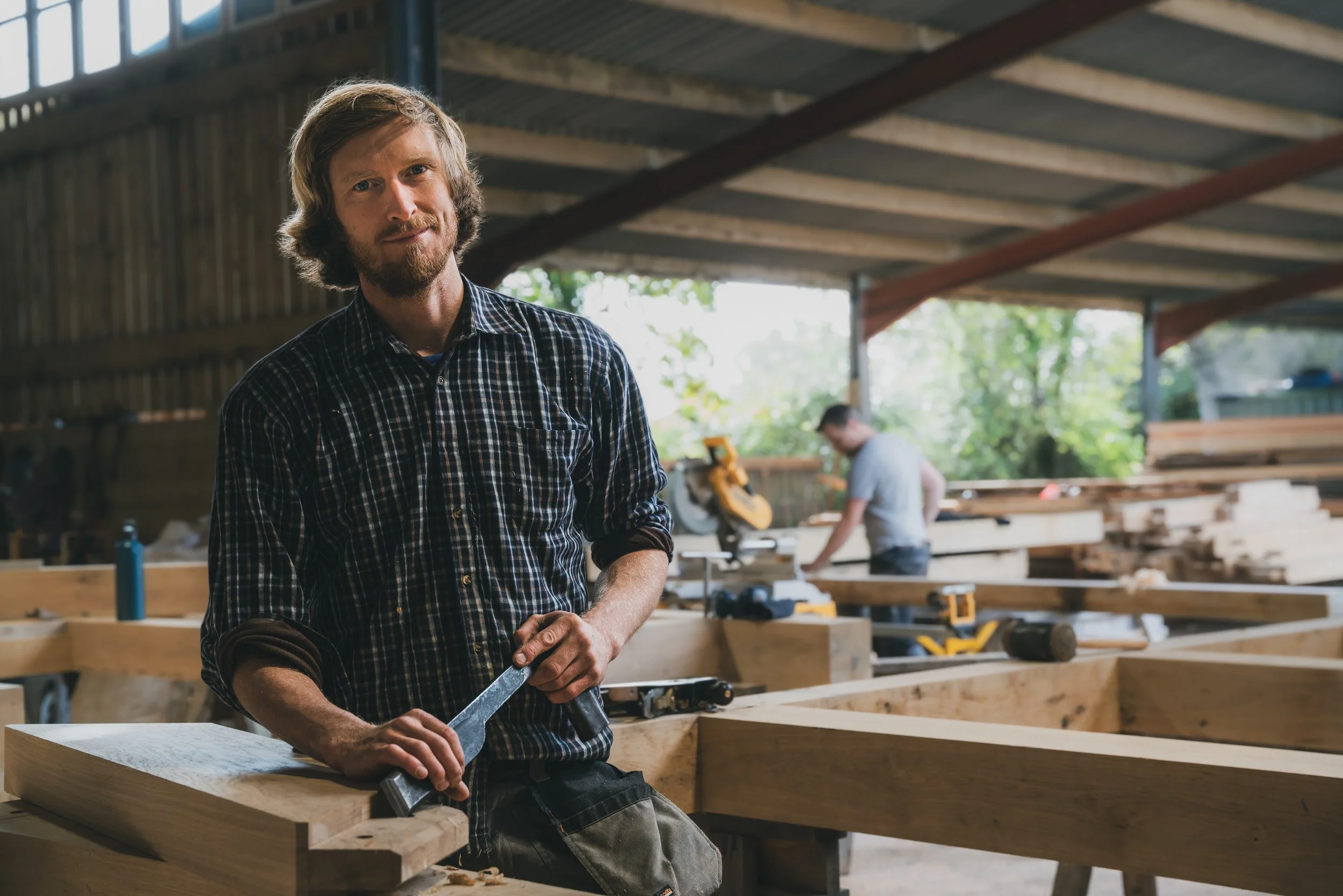 A man in a plaid shirt holding a hand saw in a woodworking workshop with another person working on wood in the background.