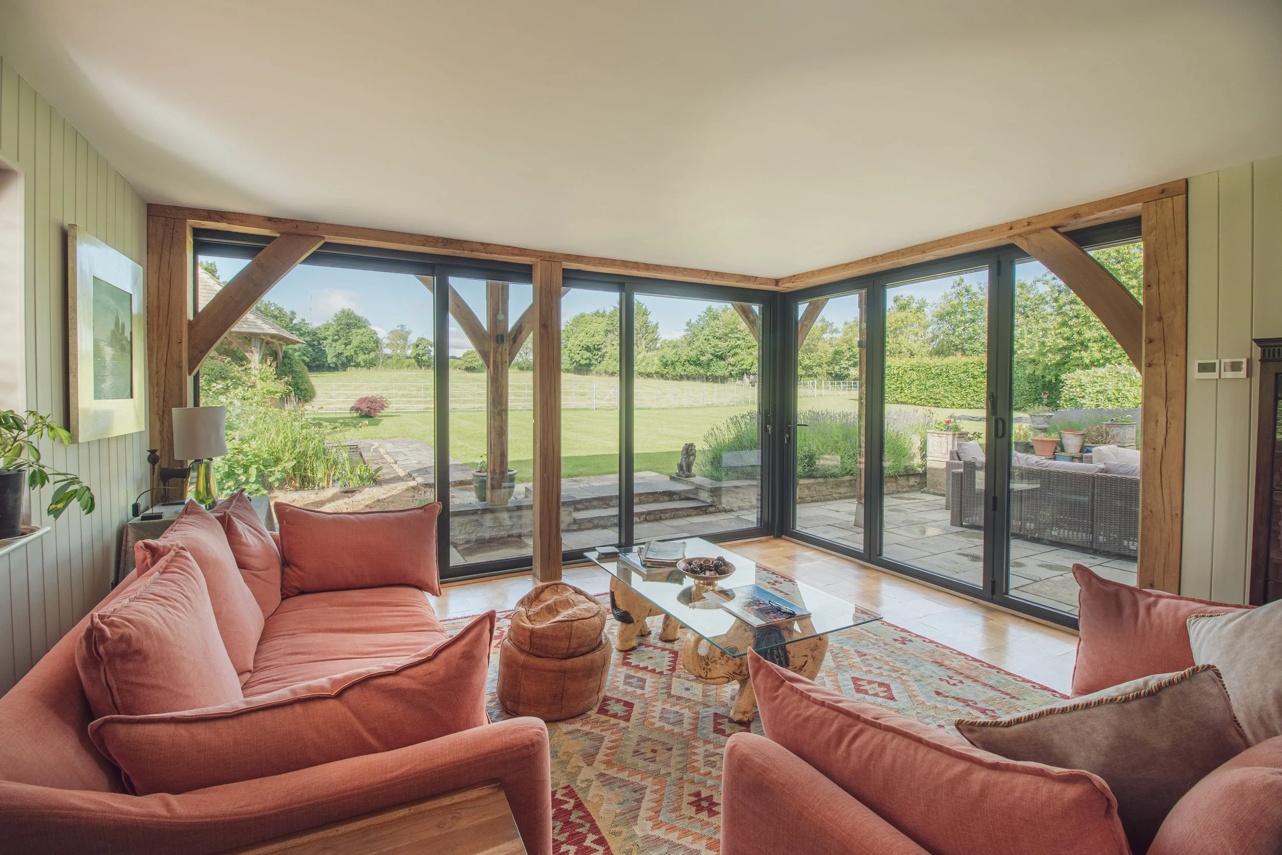 Living room with pink sofas and a glass coffee table, large glass sliding doors leading to a garden with green grass and trees, wooden beams framing the doorways, and a colorful patterned rug.