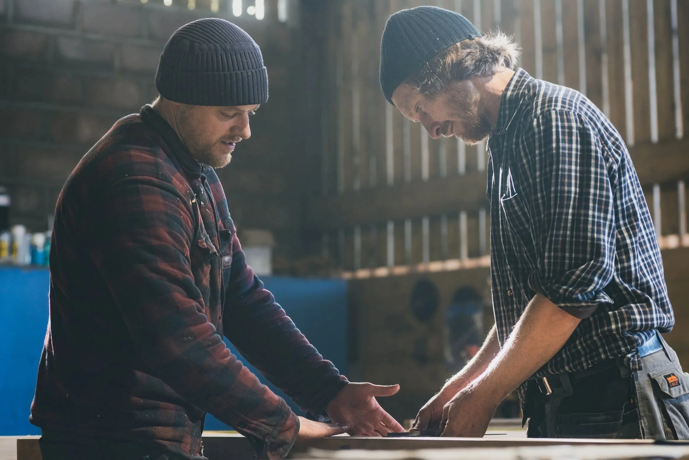 Two men in plaid shirts and beanies are working together on a woodworking project inside a workshop, looking down at the piece they are working on.