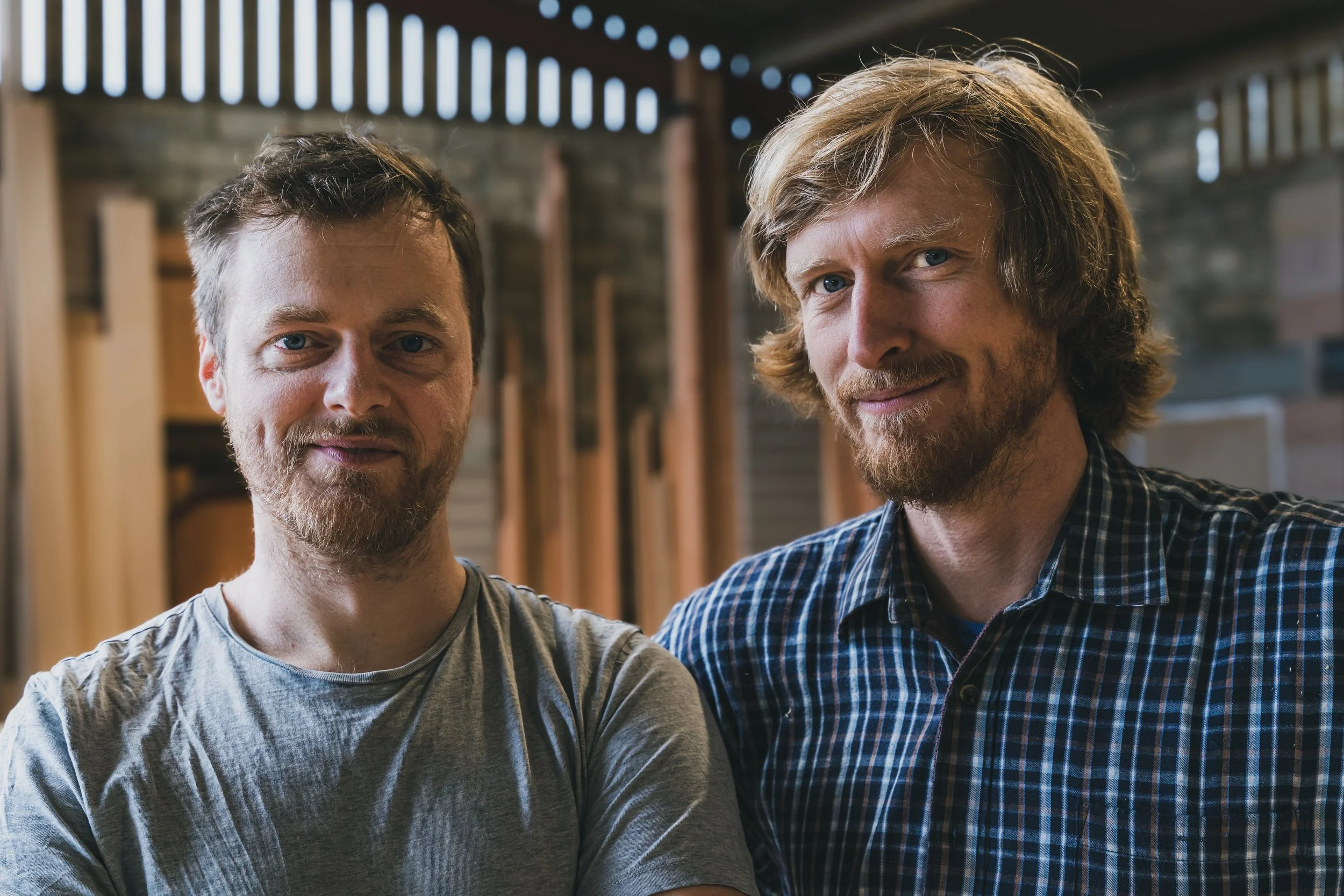 Two men standing side by side in a workshop, smiling at the camera.