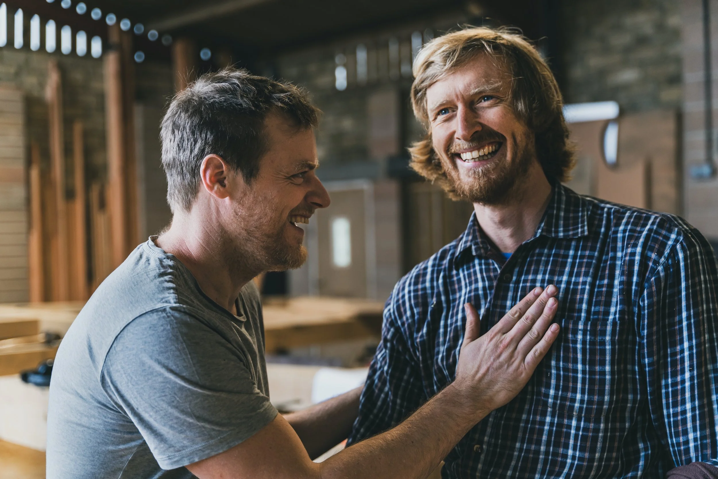 Two men smiling and laughing at each other, one touching the other's chest in an indoor setting with wooden walls.