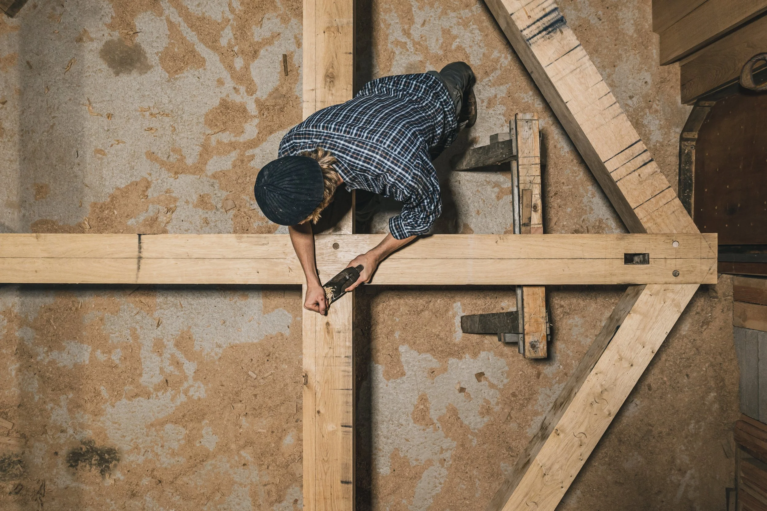 A person dressed in a plaid shirt, wearing a black beanie, is working on a wood project, using a tool on a large piece of wood, with additional wood pieces and tools nearby.