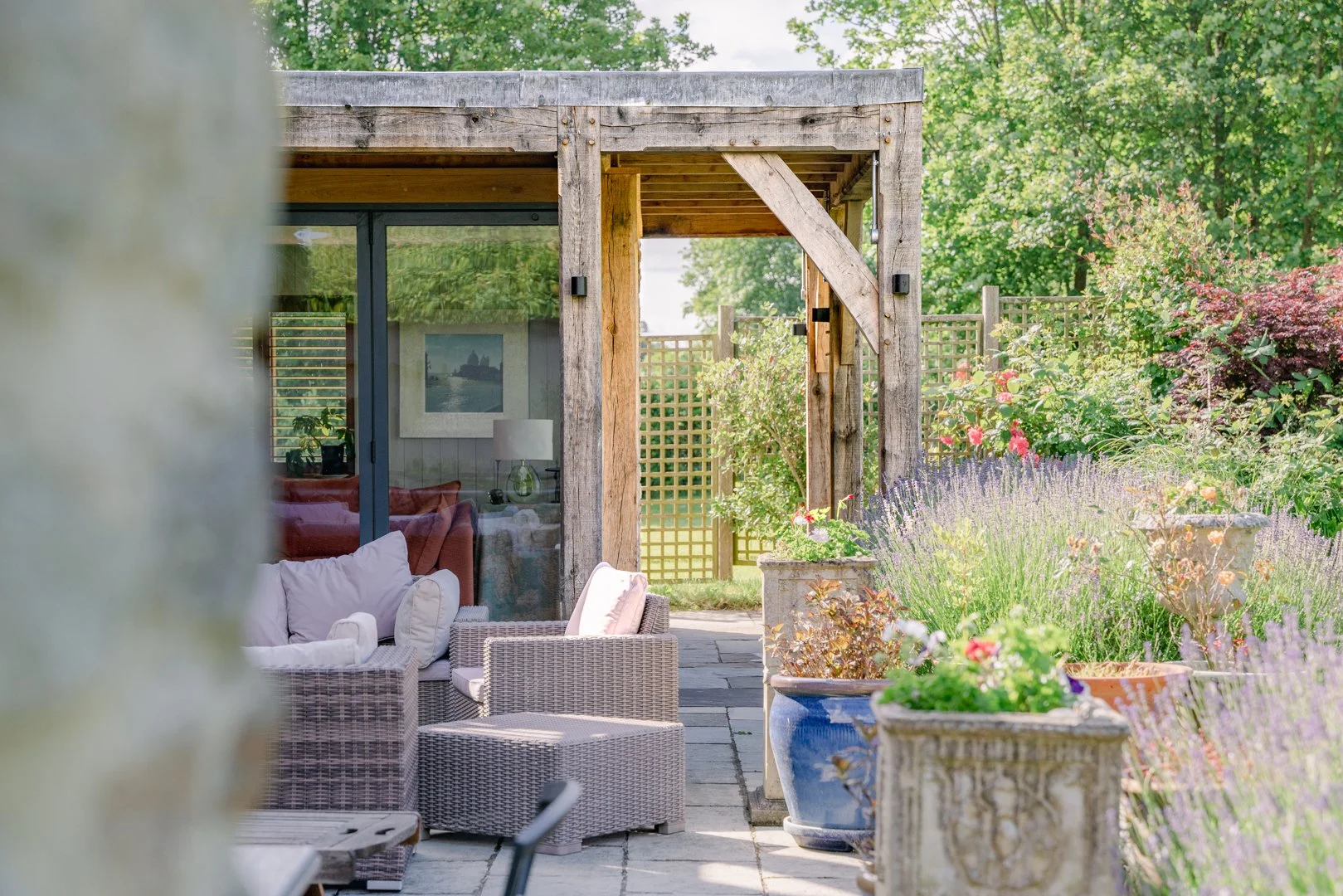 A cozy outdoor patio area with wicker furniture and throw pillows, next to a modern house with large glass windows, surrounded by lush greenery and flowering plants.