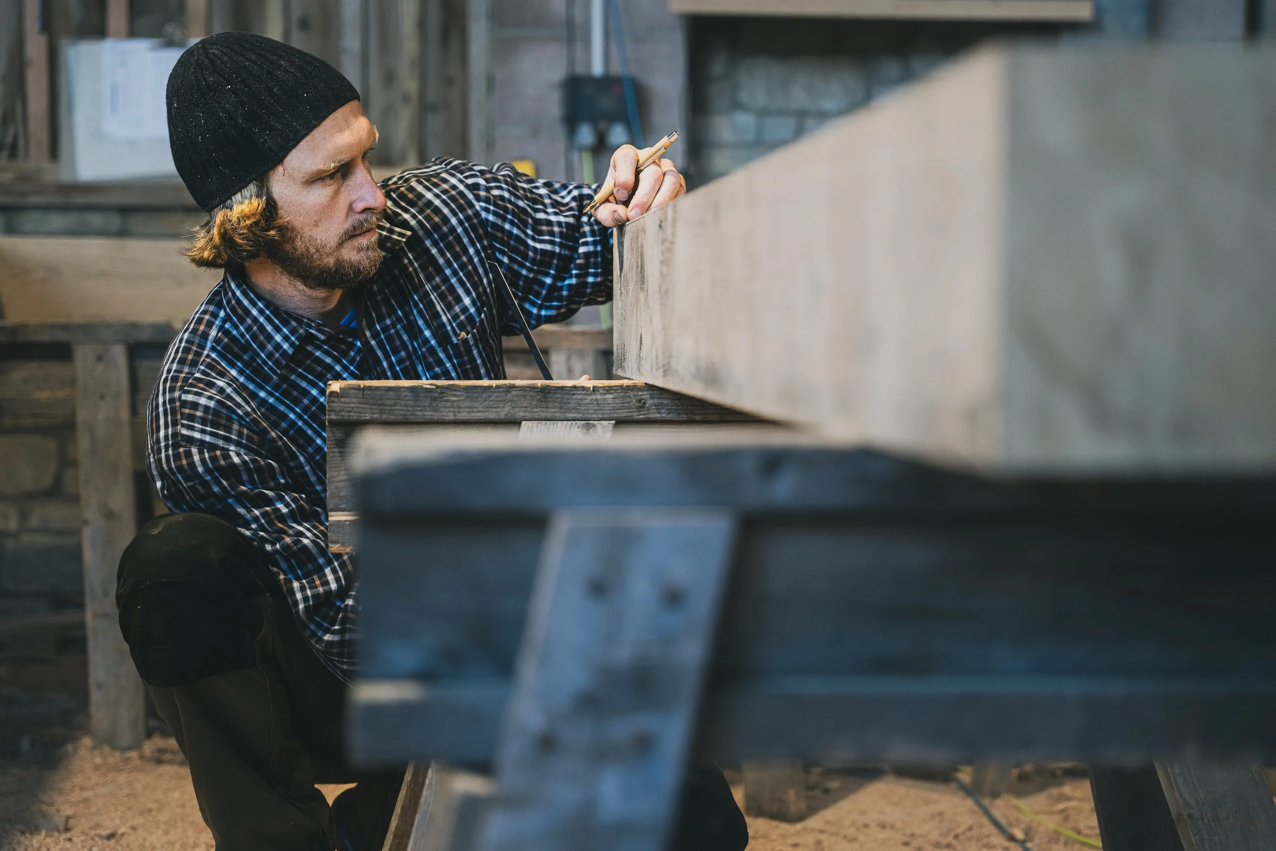 A man in a black beanie and checkered shirt work on a woodworking project in a workshop, measuring a piece of wood with a pencil.