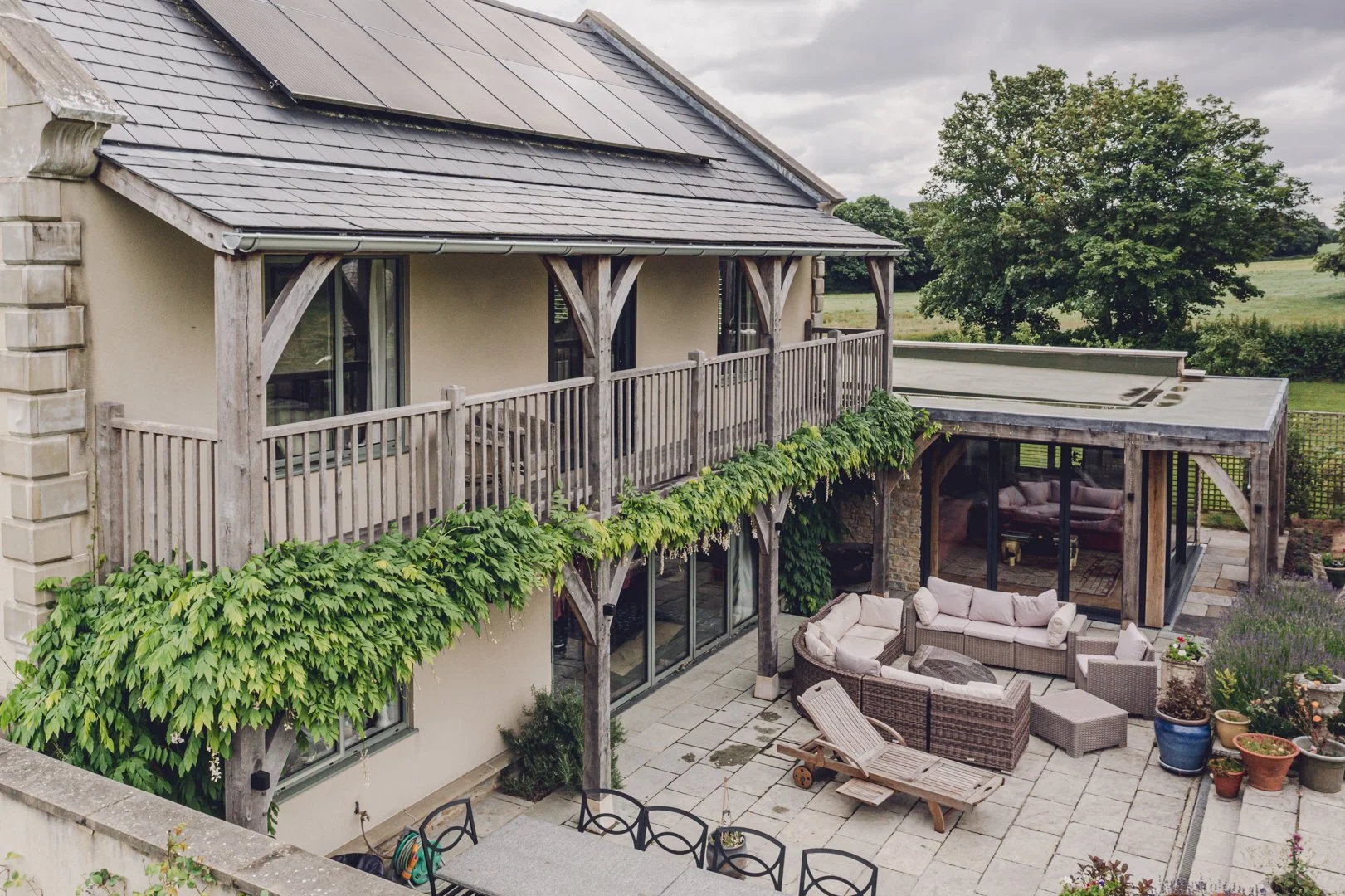 Rear view of a two-story house with a wooden balcony and glass sliding doors, outdoor patio with outdoor furniture, potted plants, and a green landscape with trees.