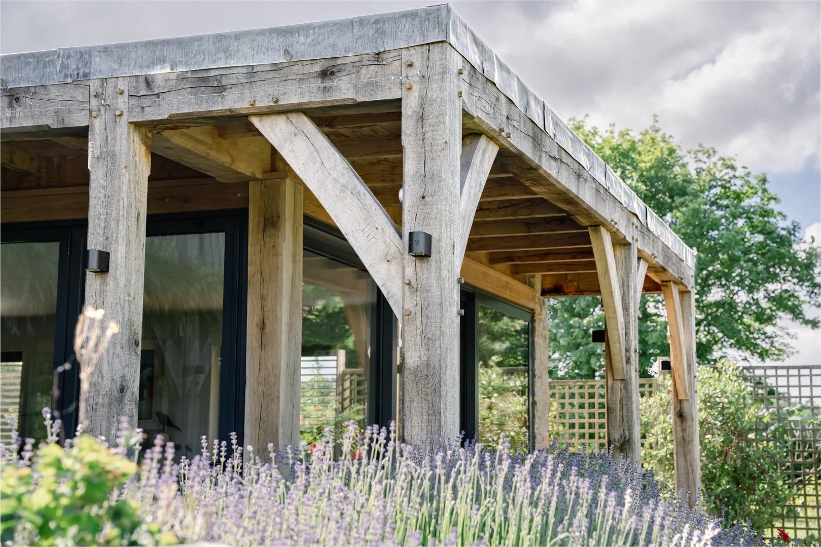A close-up view of a wooden patio cover with large beams and black mounted lights, overlooking a garden with purple lavender flowers, green foliage, and a lattice fence in the background under a partly cloudy sky.