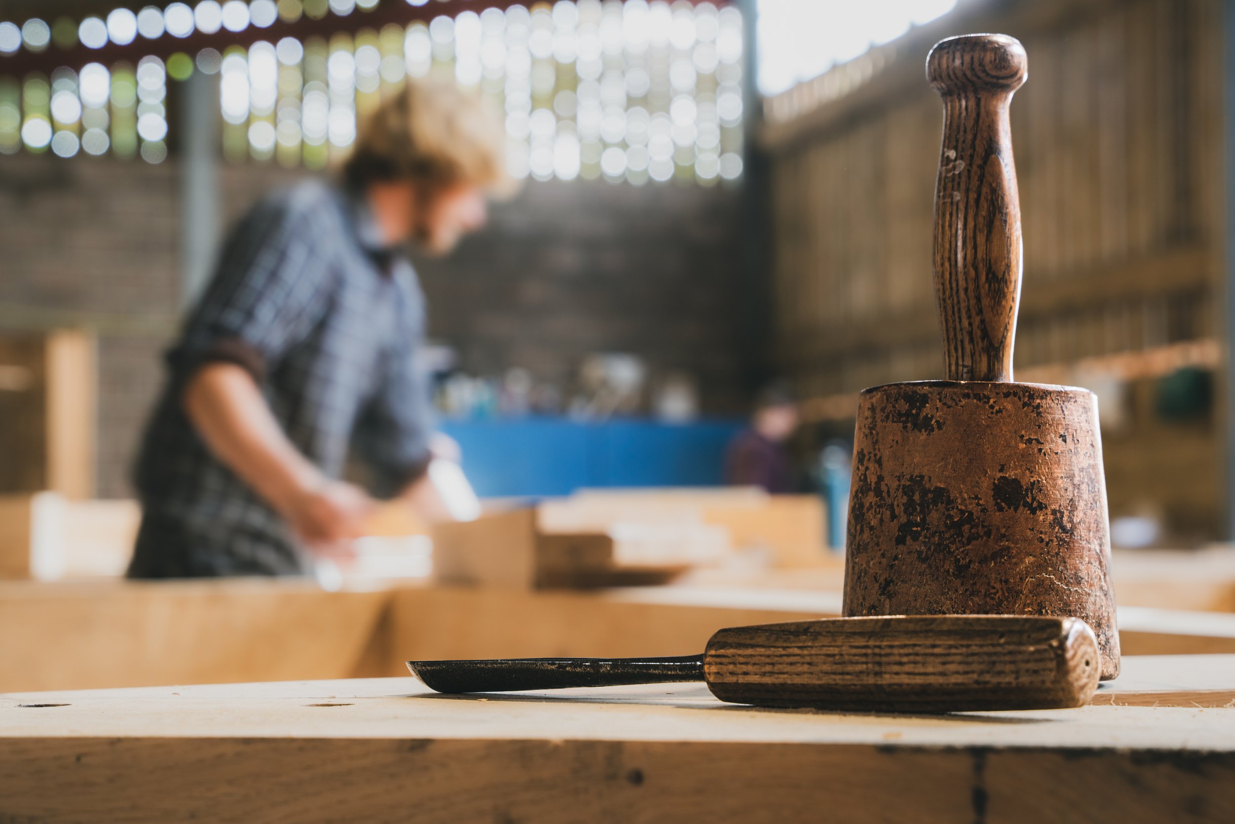 Close-up of a rusty hammer and chisel on a wooden workbench, with two blurred people working in the background inside a woodworking shop.