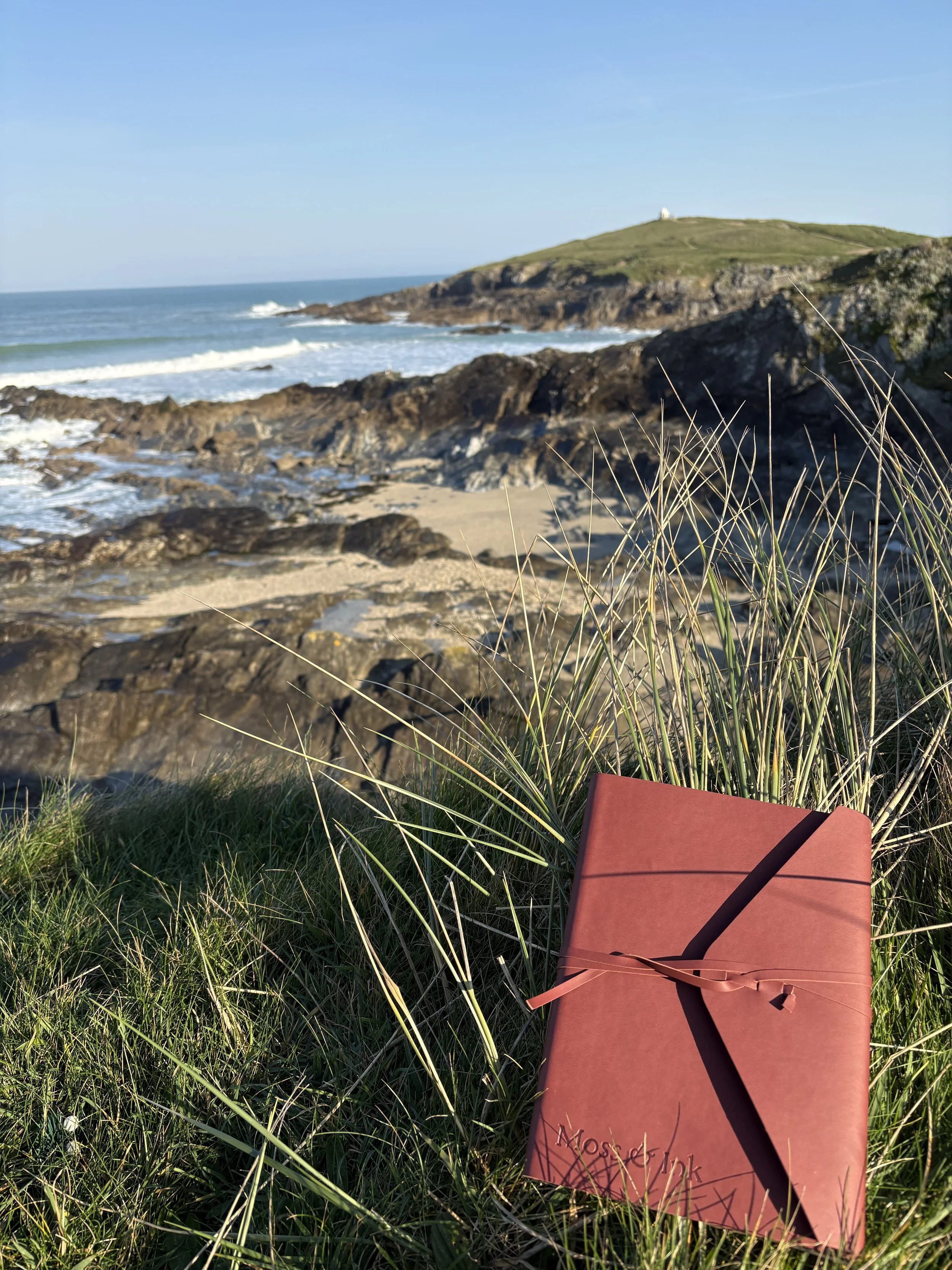 A vegan leather bound travel journal with the words 'Moss and Ink' written on it, resting on green grass near a rocky beach, with waves crashing and a hill with a structure in the distance under a clear blue sky.