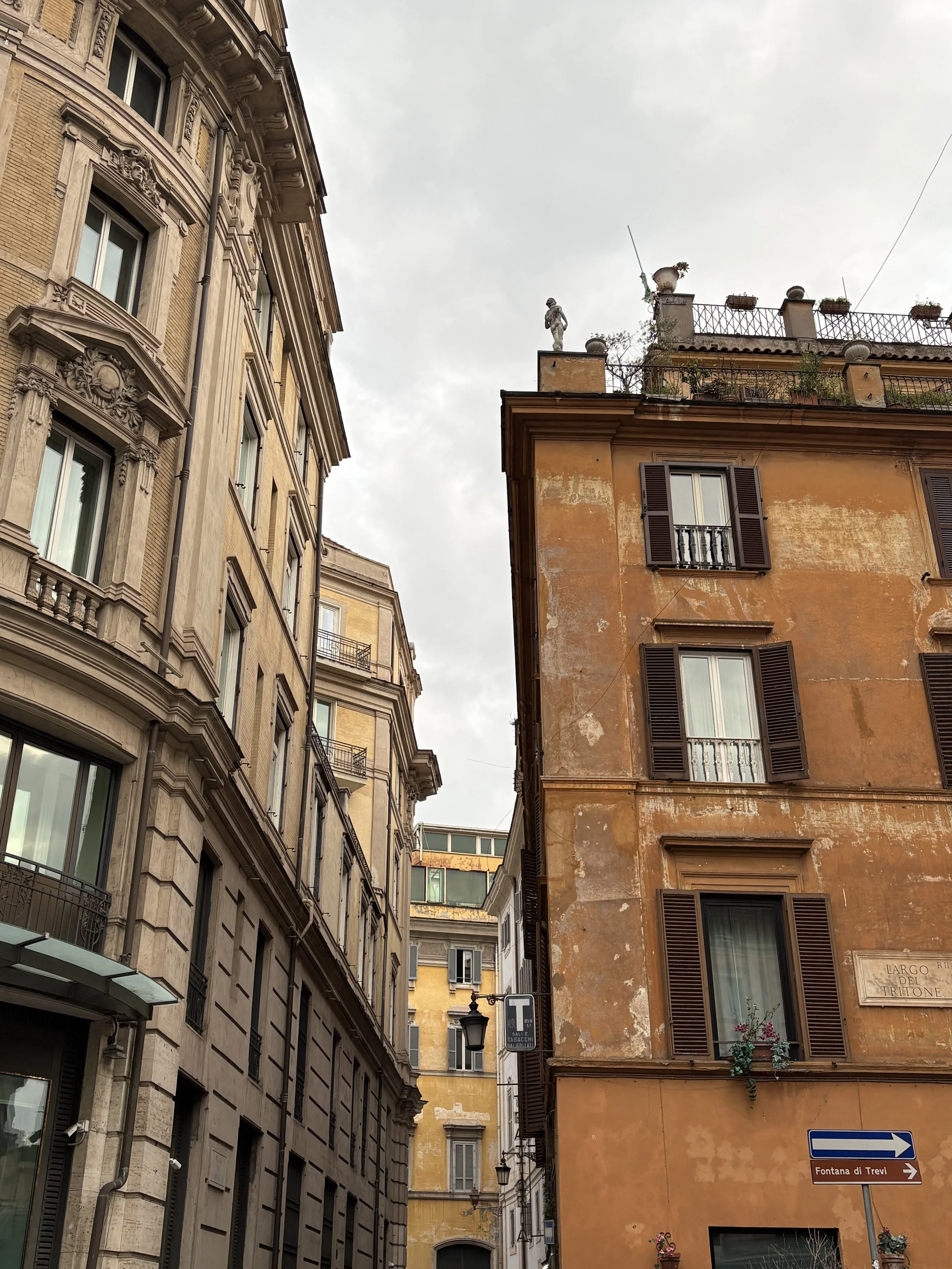 Narrow street flanked by old buildings in Rome, Italy, with street signs pointing to Fontana di Trevi and signs in Italian, under an overcast sky.