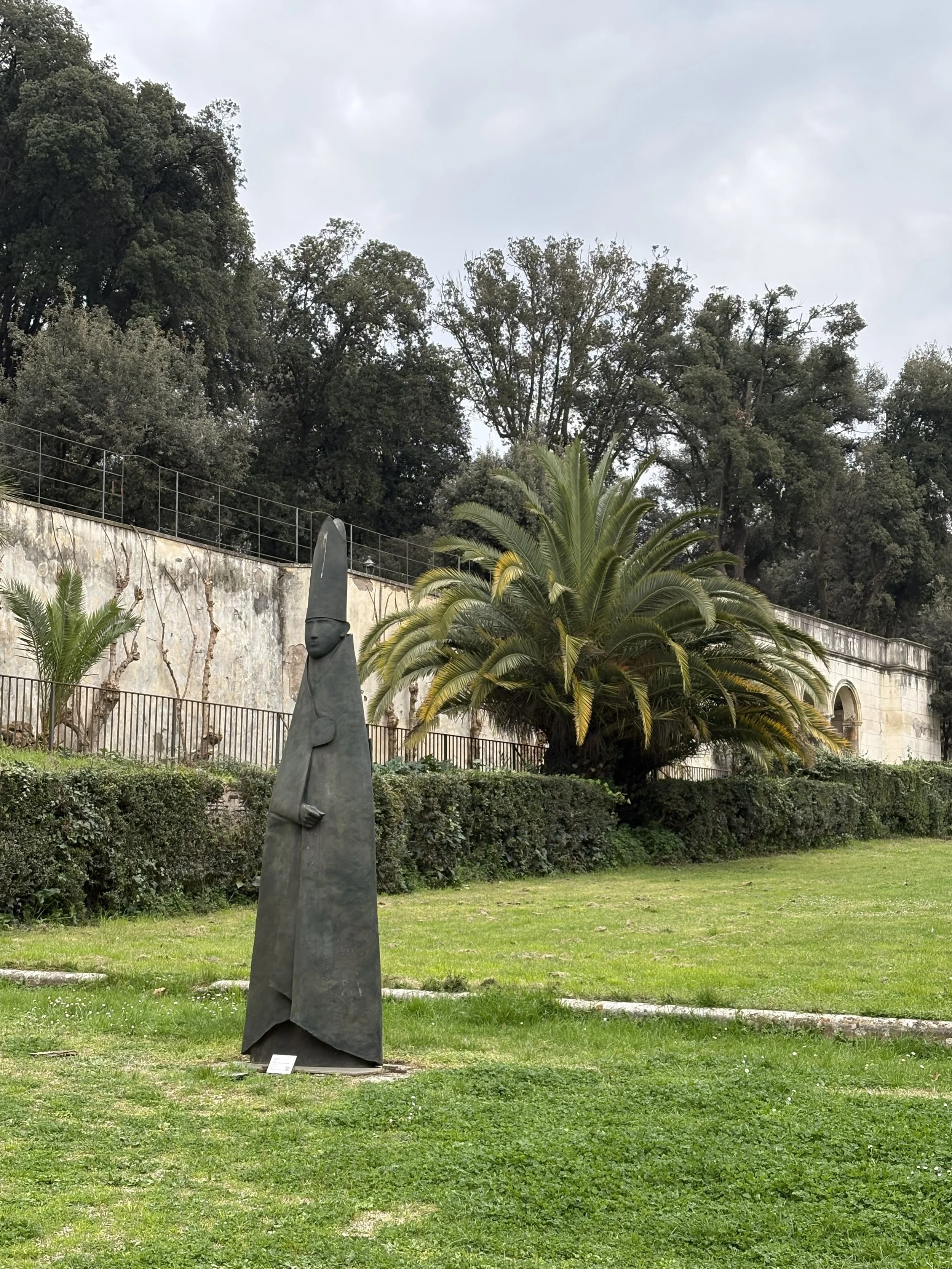 A sculpture of a tall figure in a cloak with a headpiece, set outdoors on a grassy area with bushes and large trees in the background.