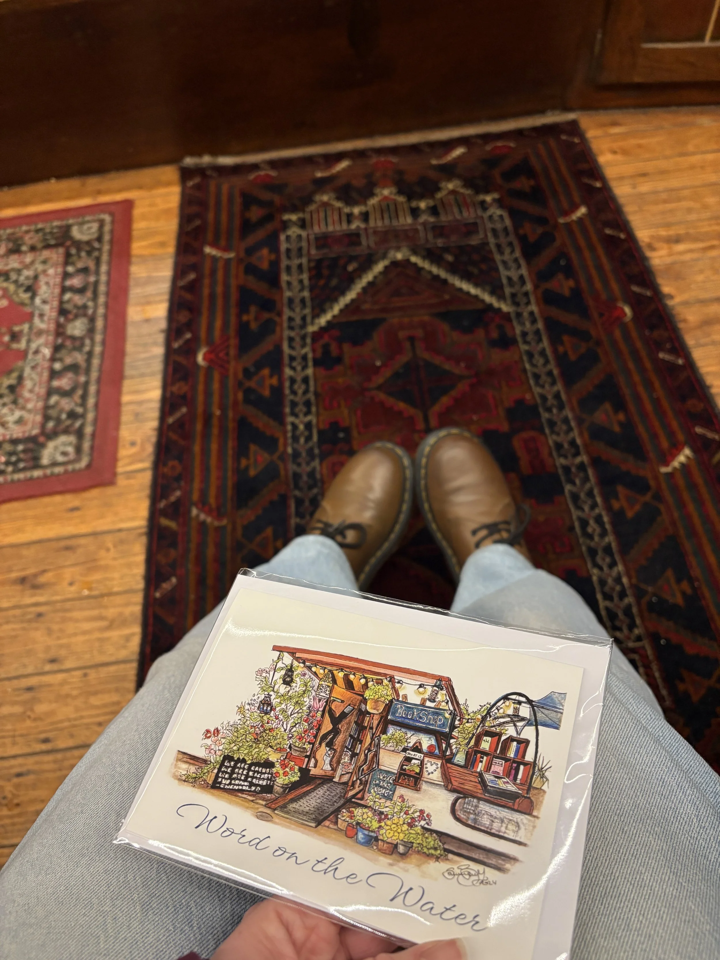 A person wearing blue jeans and brown boots holding a colourful illustrated brochure or booklet titled 'Wood on the Water' with a watercolor building facade, potted plants, and sign 'Book Shop' in front of a wooden floor and rugs.