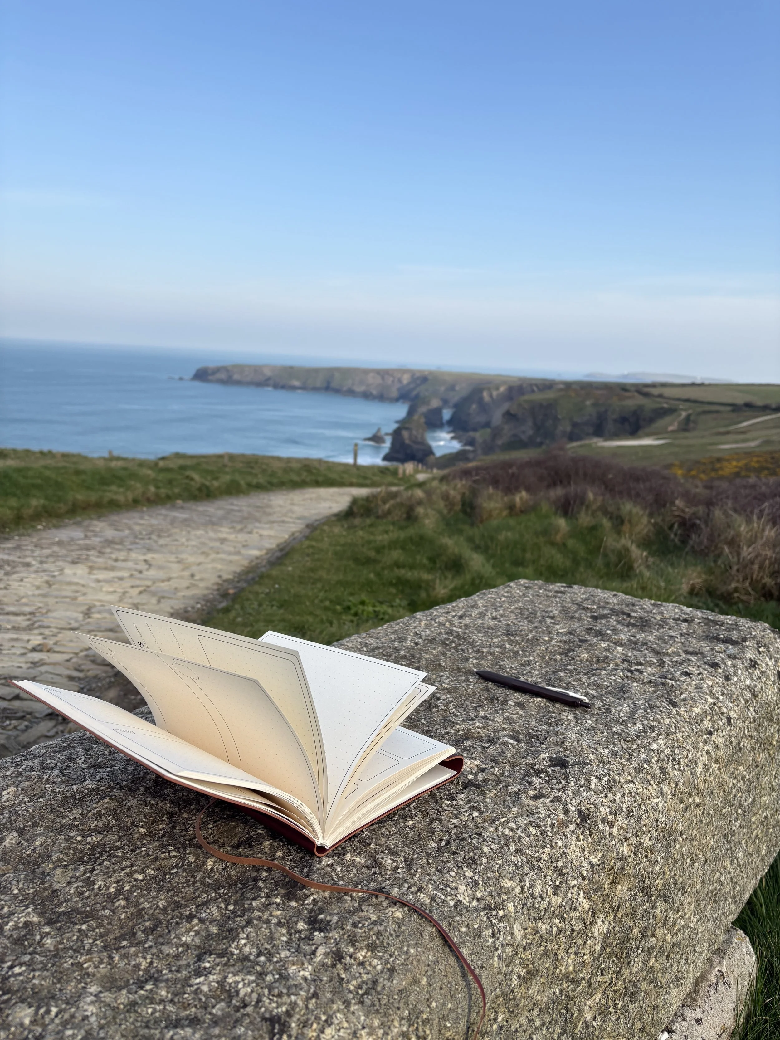 Open notebook and pen on a large stone with a scenic coastal cliff view in the background.