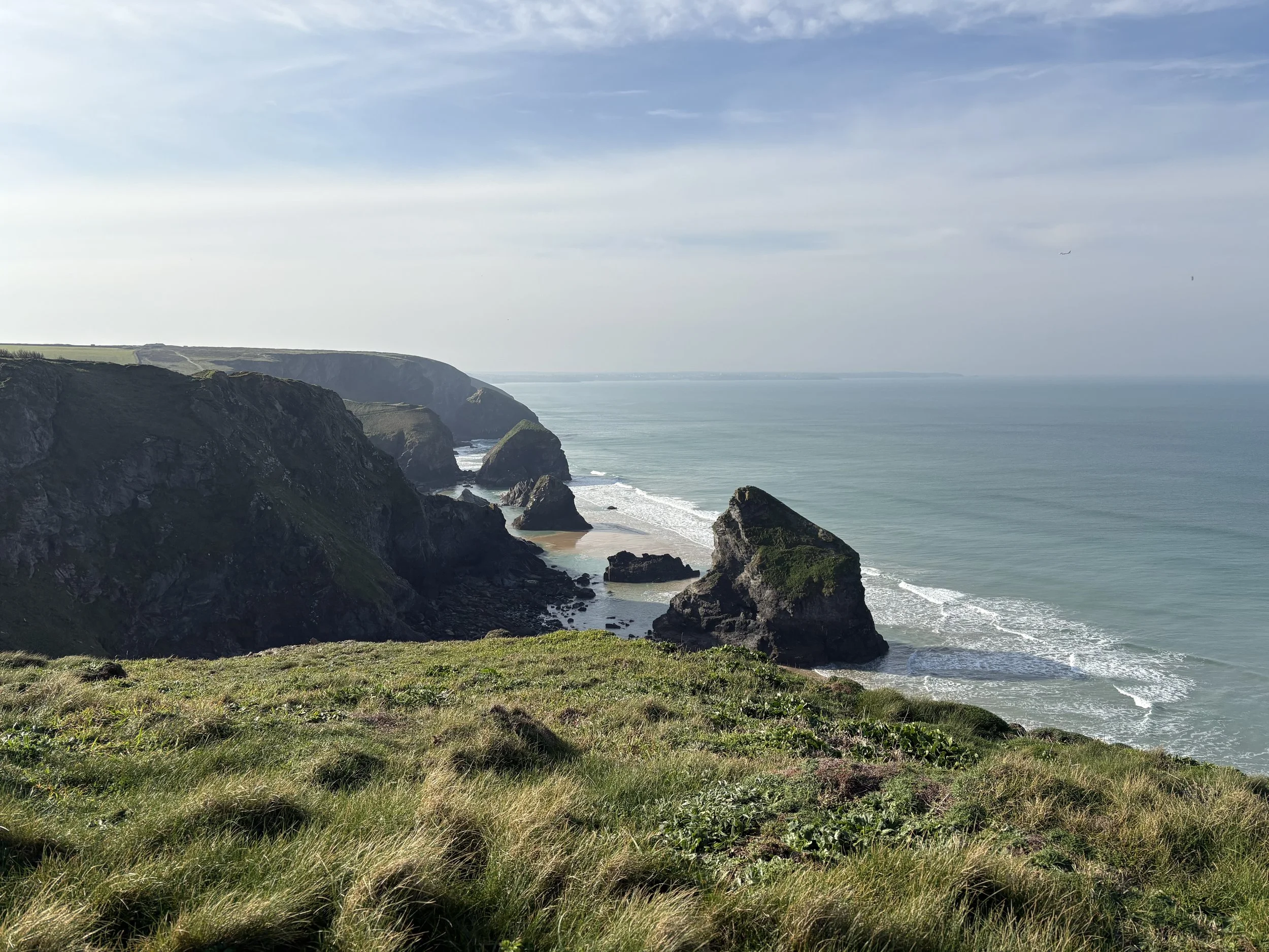 Coastal cliffs with green grass on top, rocky formations extending into the ocean, and a sandy beach below. The ocean is calm with gentle waves, under a partly cloudy sky with a few airplanes visible.