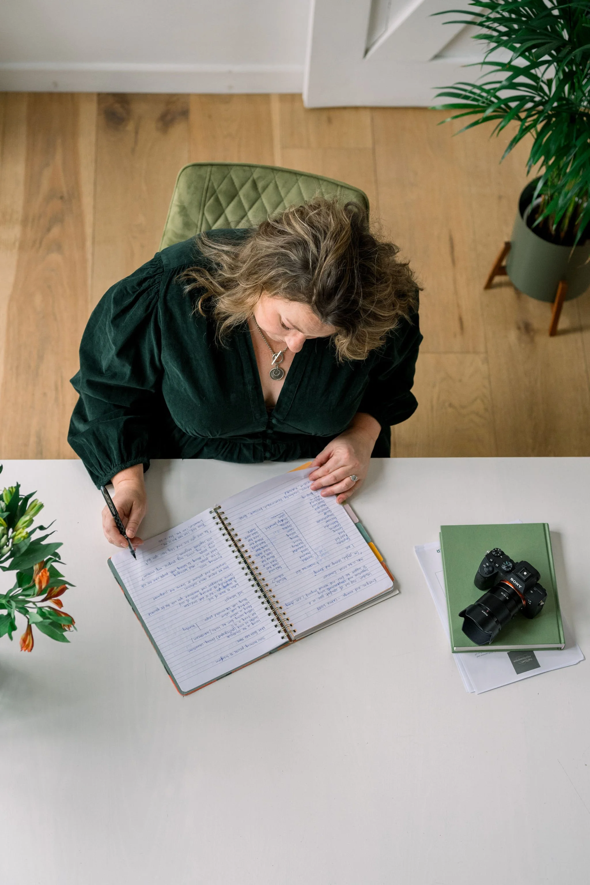 Top-down view of a woman with curly hair in a black blouse, sitting at a white desk, writing in a spiral notebook, with a camera and green books on her right, and a potted plant on her left.