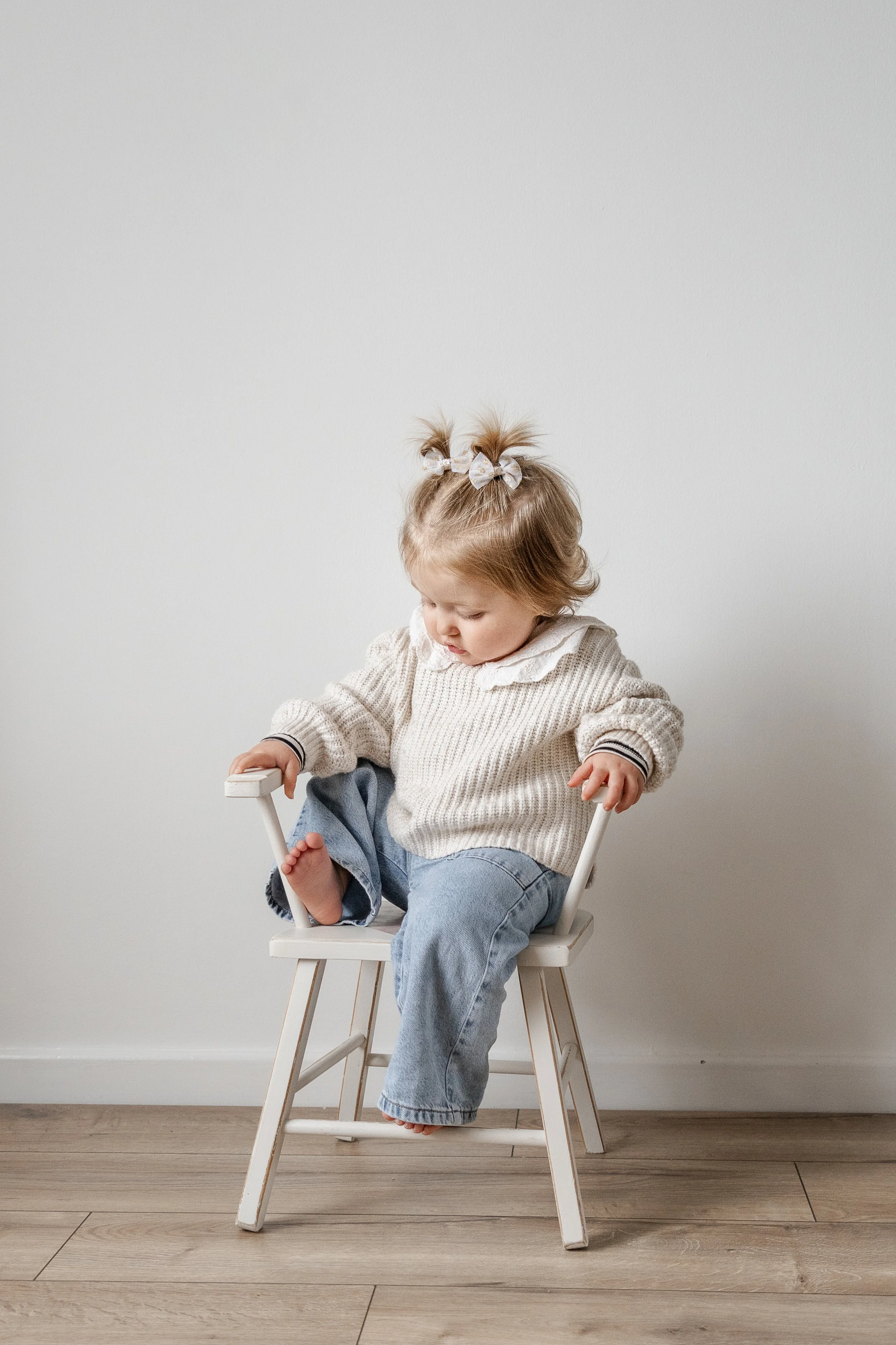 Young girl sitting on a small white chair against a plain white wall.