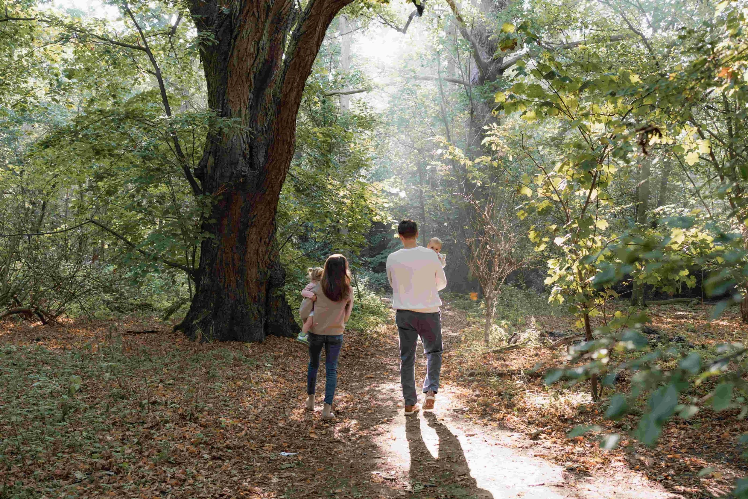 A family of four walking through a wooded forest trail during daylight. The father is holding a small child, the mother walking behind with a young girl, and the son walking slightly apart in front of the others. Sunlight filters through the green le