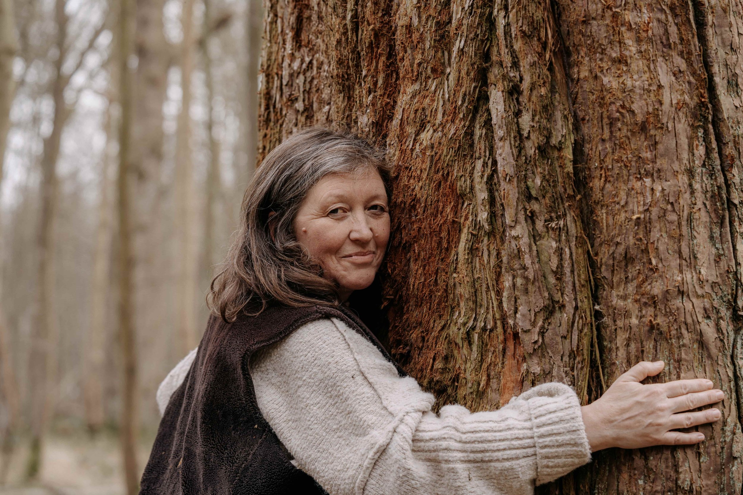 A woman with long brown hair hugging a large tree in a forest.