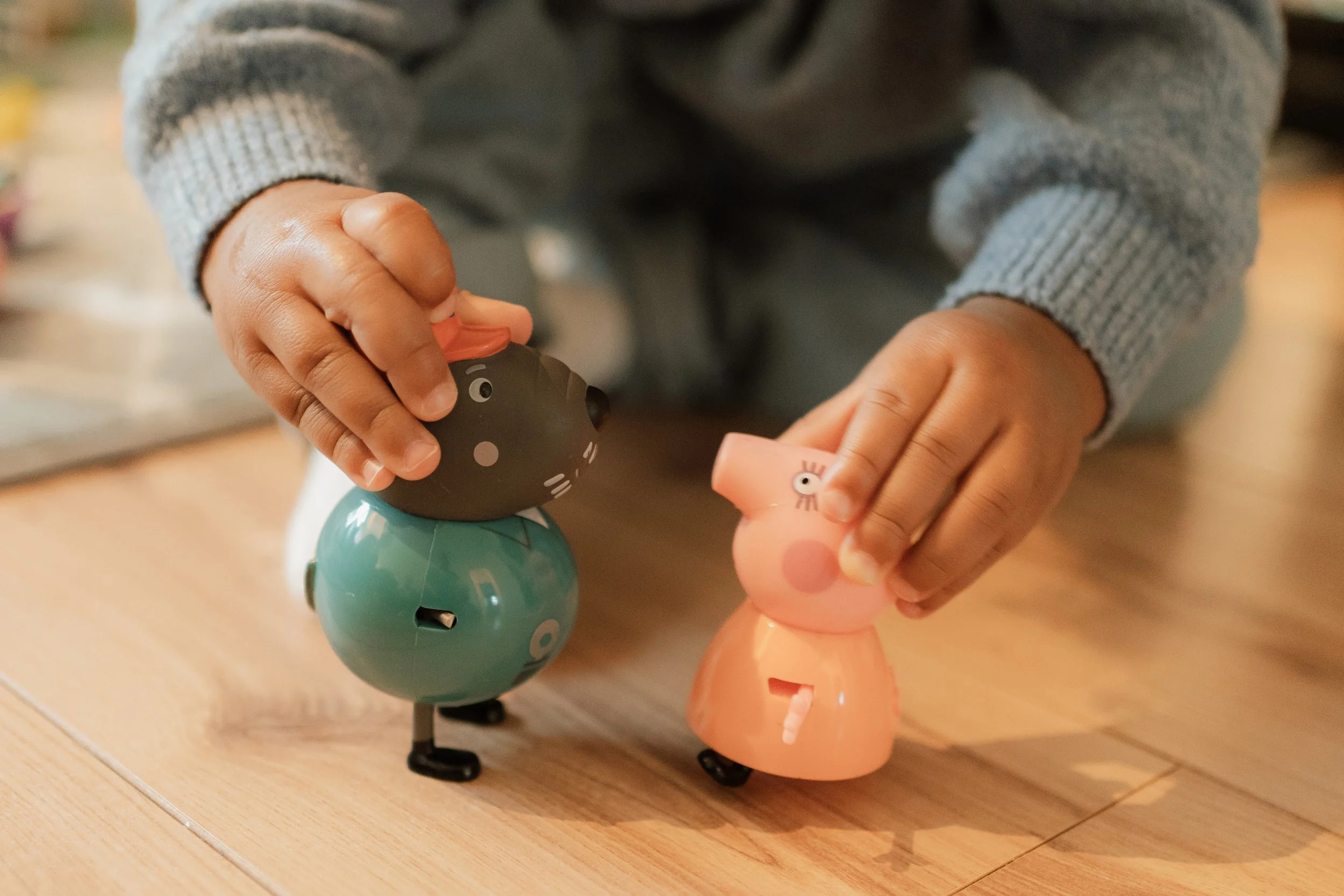 A child playing with Peppa Pig and Harry Potter coin banks on a wooden floor, raising the coin banks in their hands.