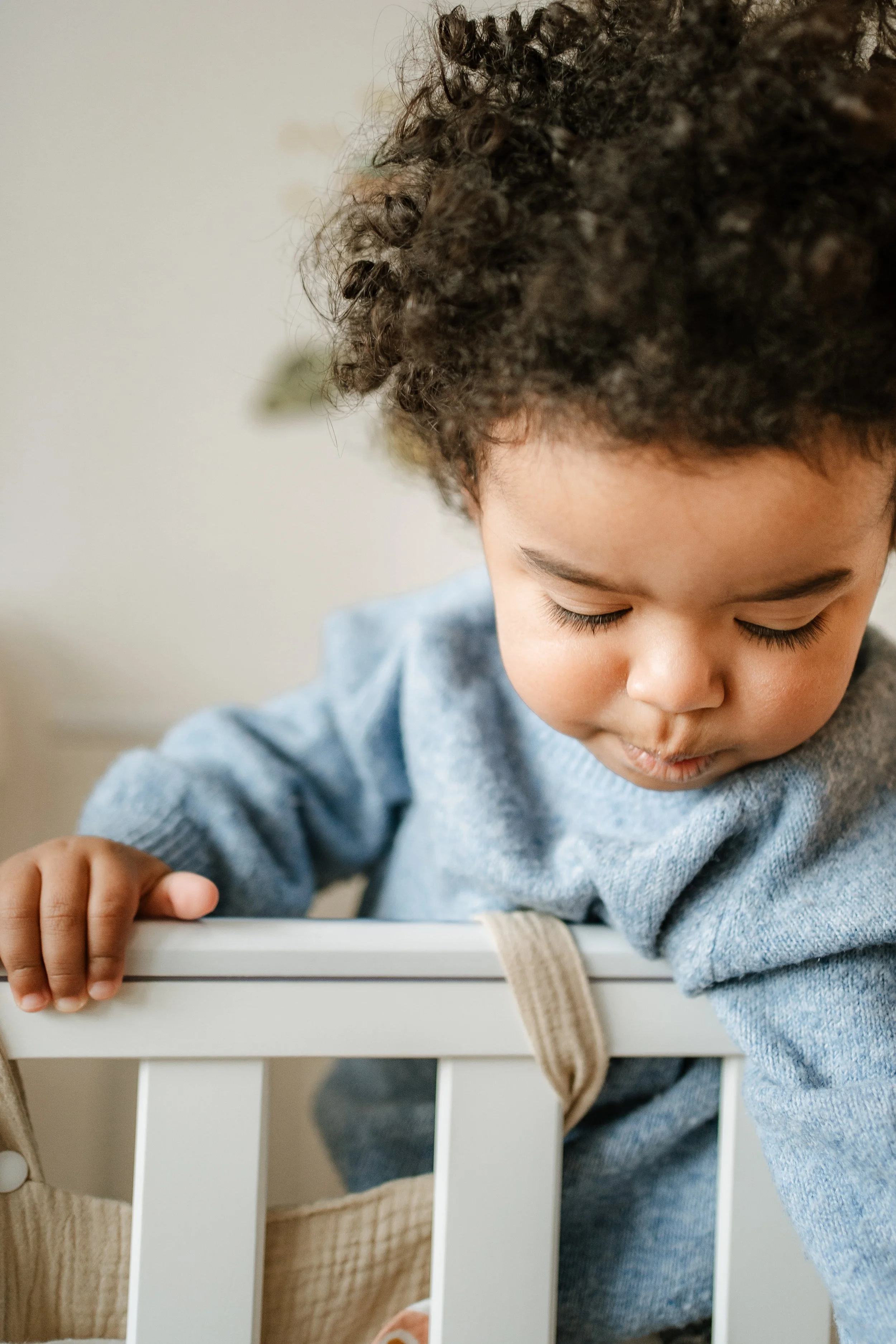 Young child leaning over the side of a white crib, looking downwards with curly hair and wearing a blue sweater.