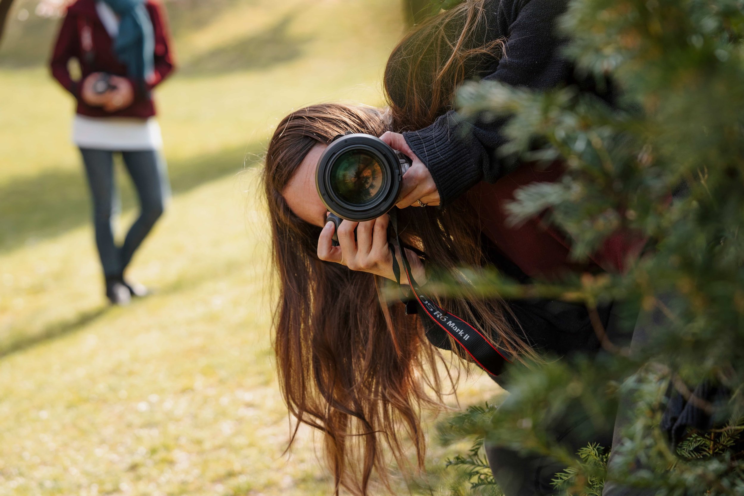 A woman with long brown hair taking a photograph outdoors with a camera, leaning over a bush, with a person in a red jacket and gray pants standing in the background.