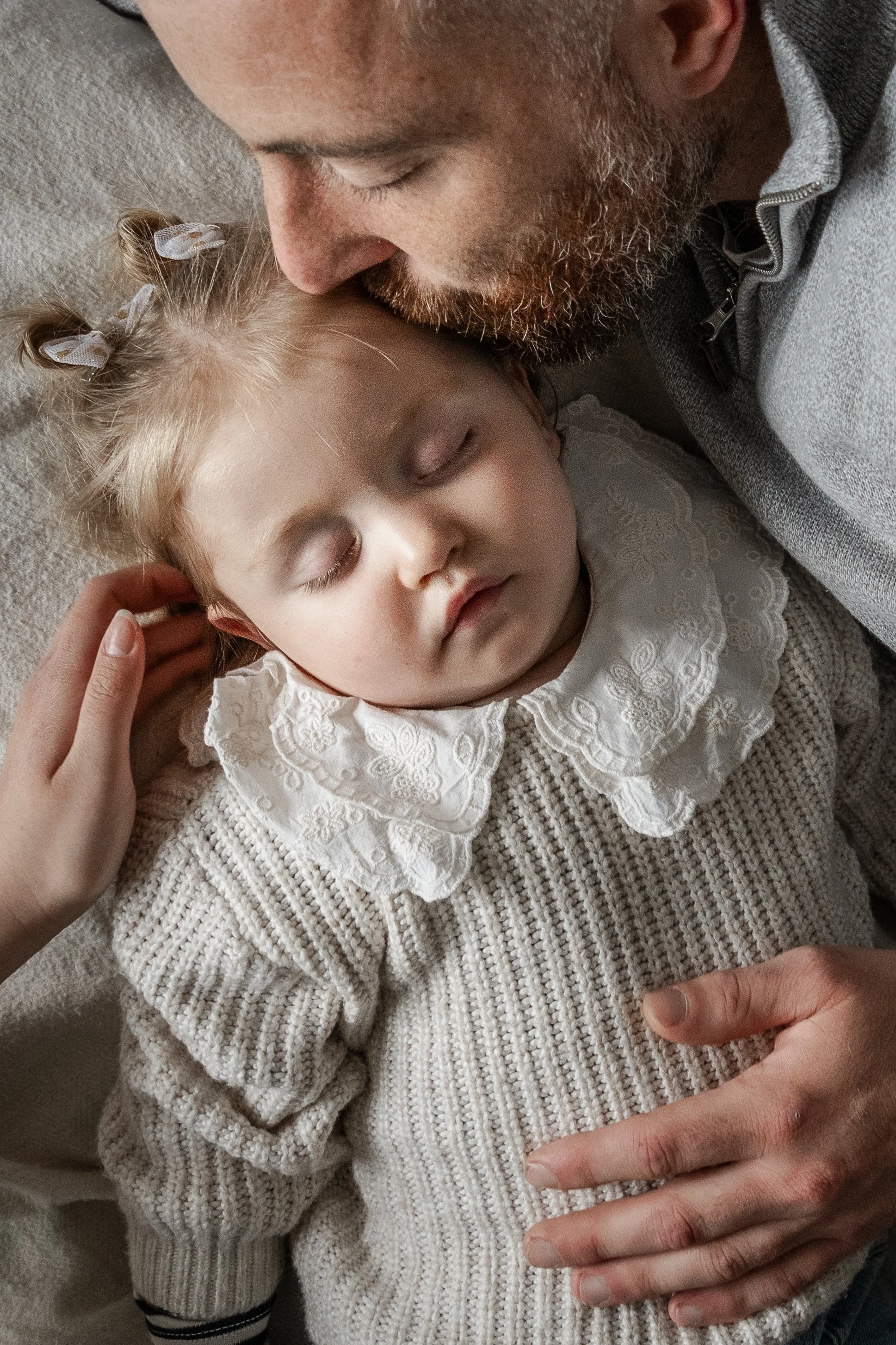 A man with a beard and a gray zip-up jacket is gently kissing a sleeping young girl with blonde hair styled in two pigtails tied with white bows, dressed in a cream-colored sweater with a white embroidered collar, as she lies on the floor or a soft s