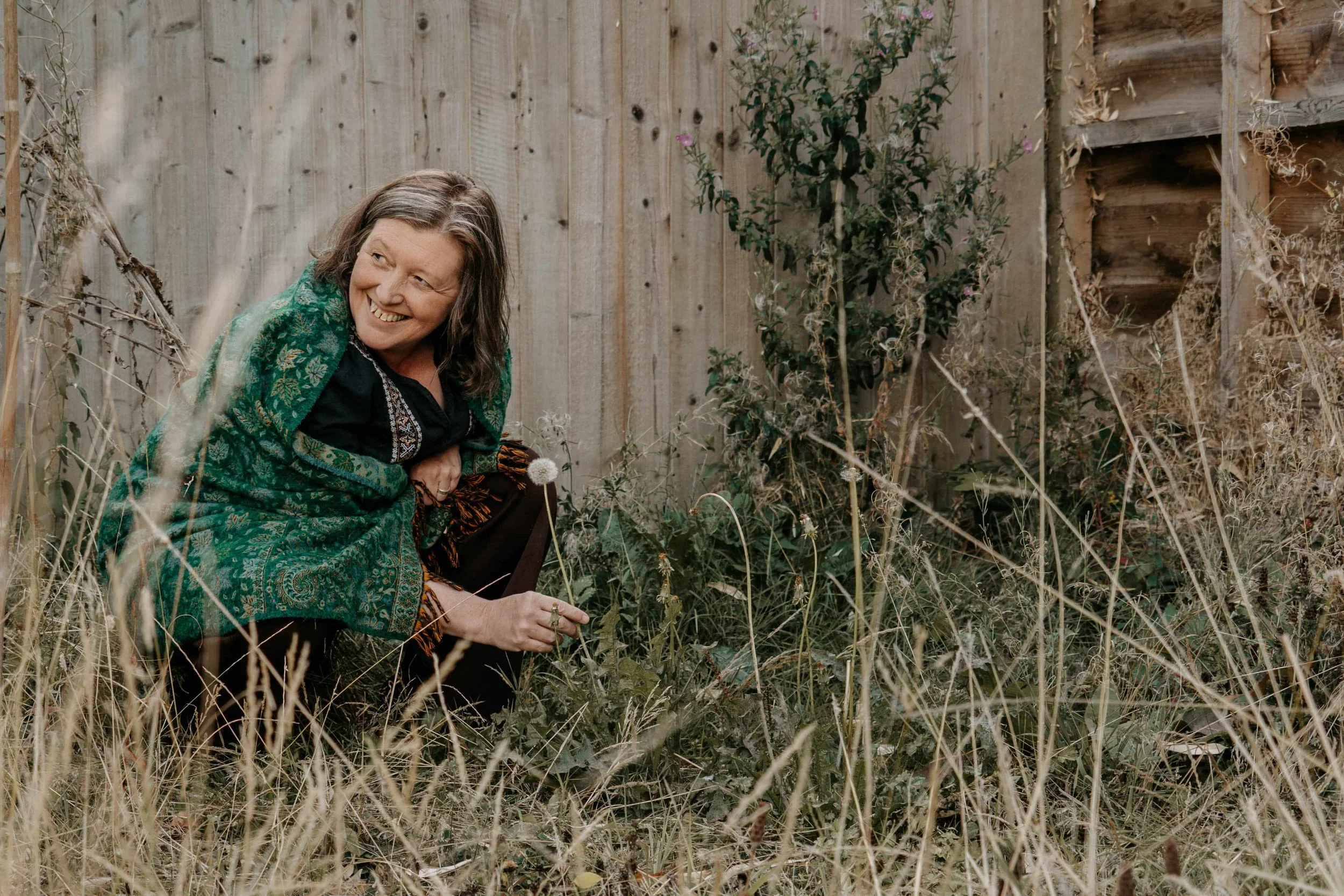 A woman with brown hair smiling, wearing a black shirt and a green patterned shawl, kneeling in overgrown grass and weeds in a backyard or garden, with a wooden fence in the background.