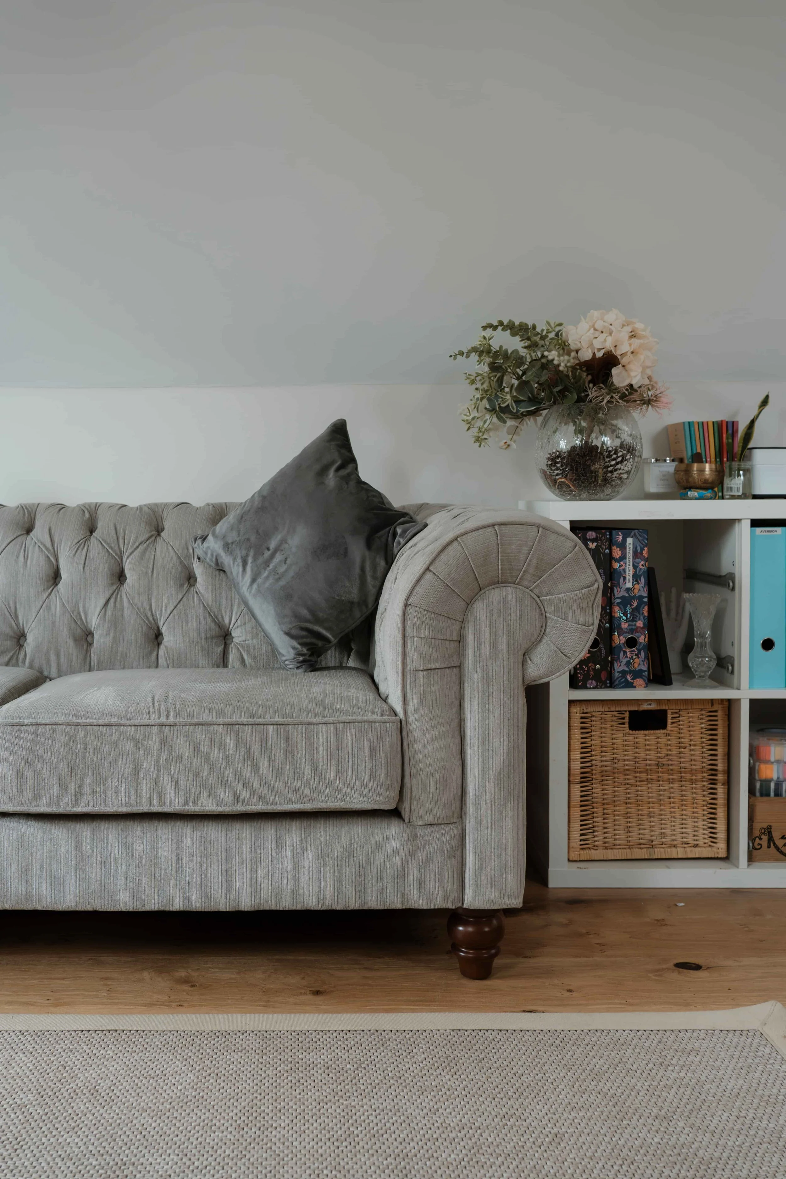 A beige tufted sofa with a dark gray pillow, next to a white bookshelf with books, decorative items, and a large glass vase with flowers, all on a hardwood floor.