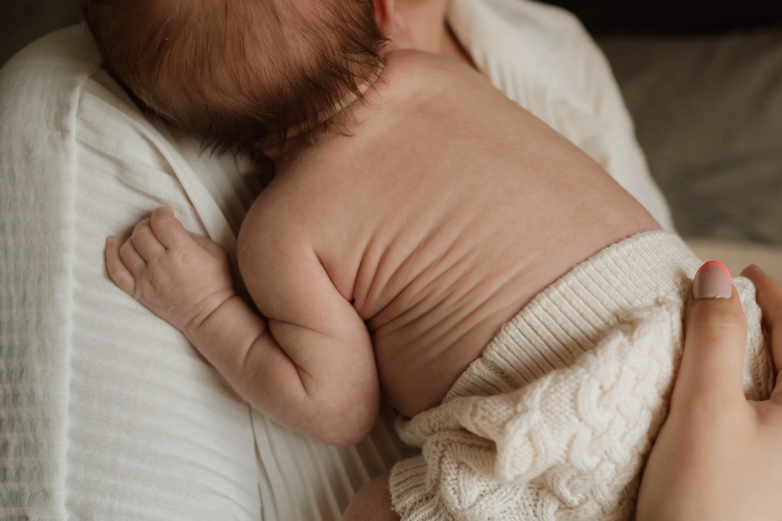 Close-up of a newborn baby breastfeeding while lying on a person's chest, with the person's hand gently supporting the baby's back.