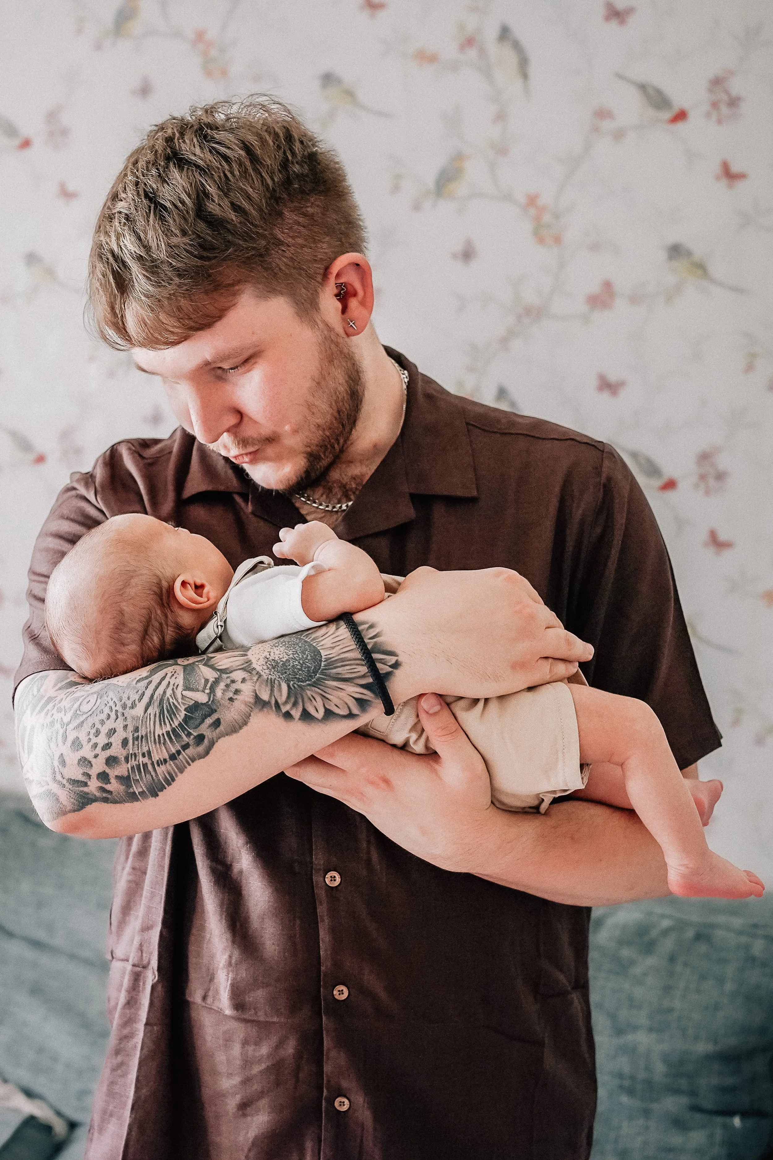 A young man with tattoos on his arm holding a baby in his arms, looking down at the baby affectionately in a room with bird-patterned wallpaper.