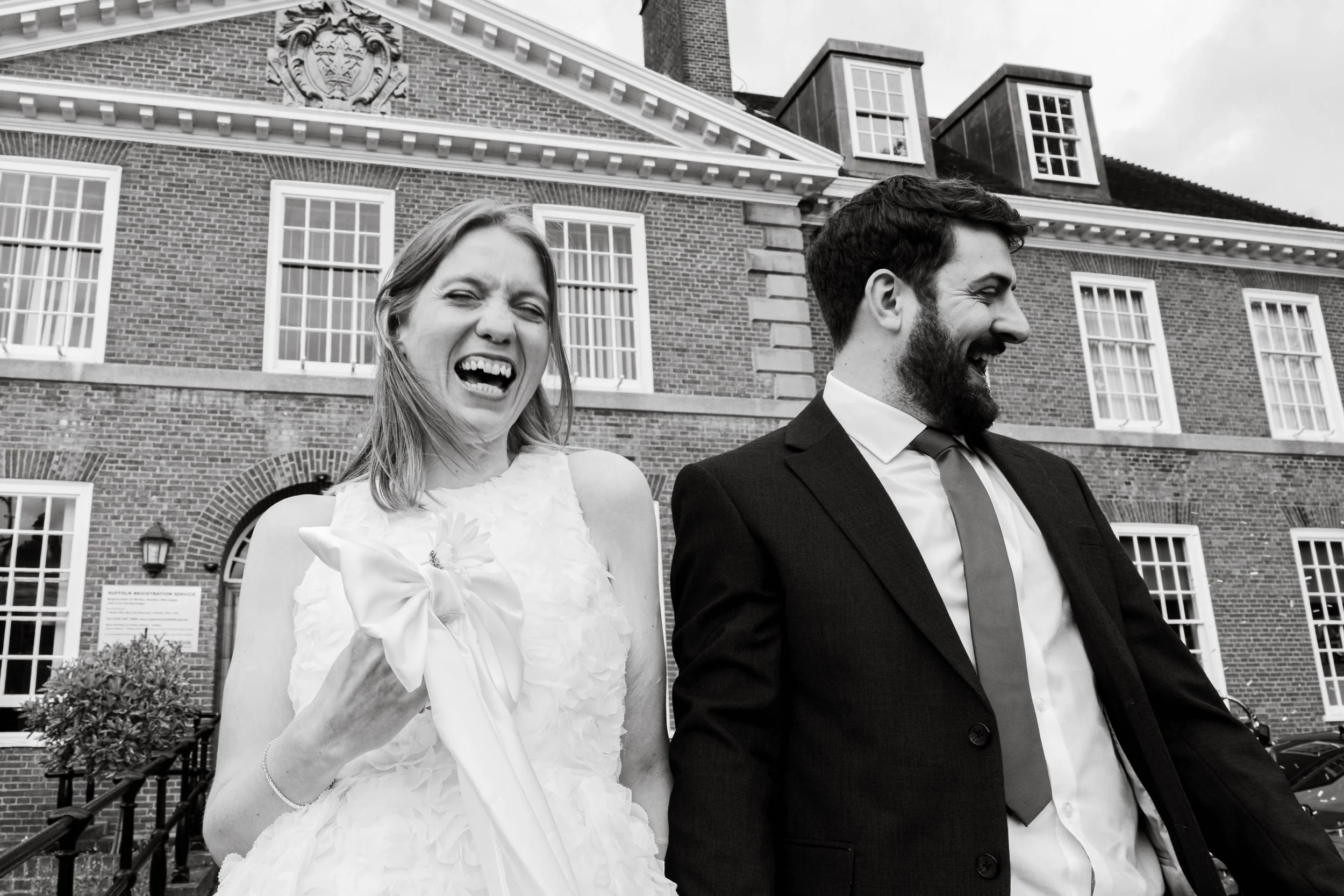 A woman and man in formal attire are laughing outdoors in front of a large brick building. The woman is holding a bouquet and wearing a wedding dress, and the man is in a suit with a tie.