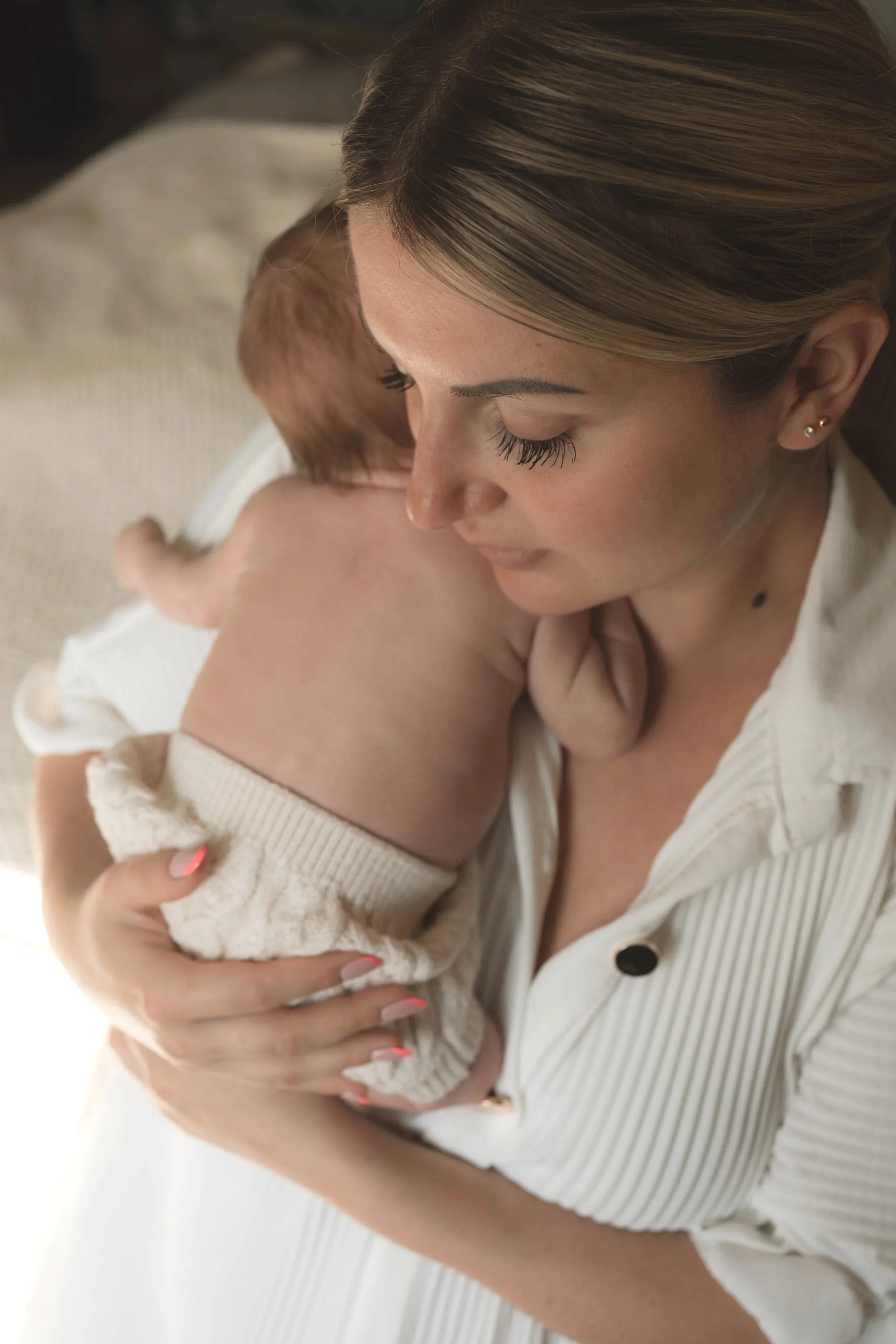 A woman with blonde hair holding and cuddling a newborn baby with reddish hair, both dressed in neutral-colored clothes, in a close-up indoor setting.
