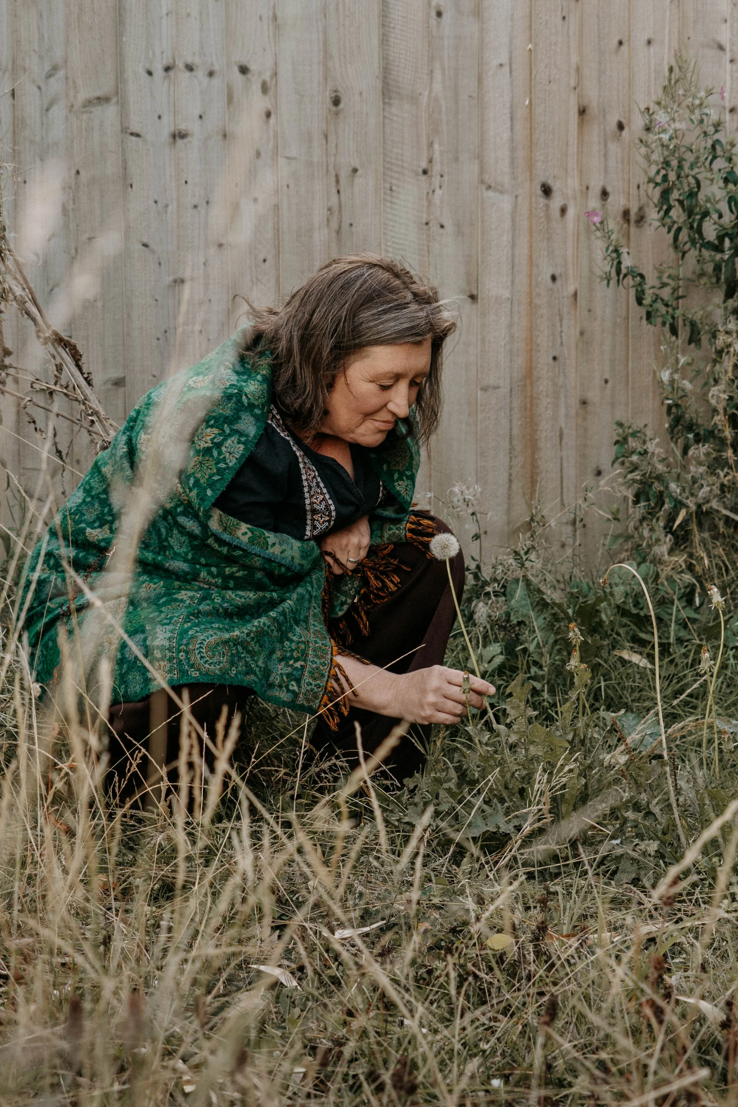 A woman with gray hair wearing a black top and green shawl with intricate patterns, crouching in a grassy backyard with a wooden fence in the background, holding a dandelion seed head.