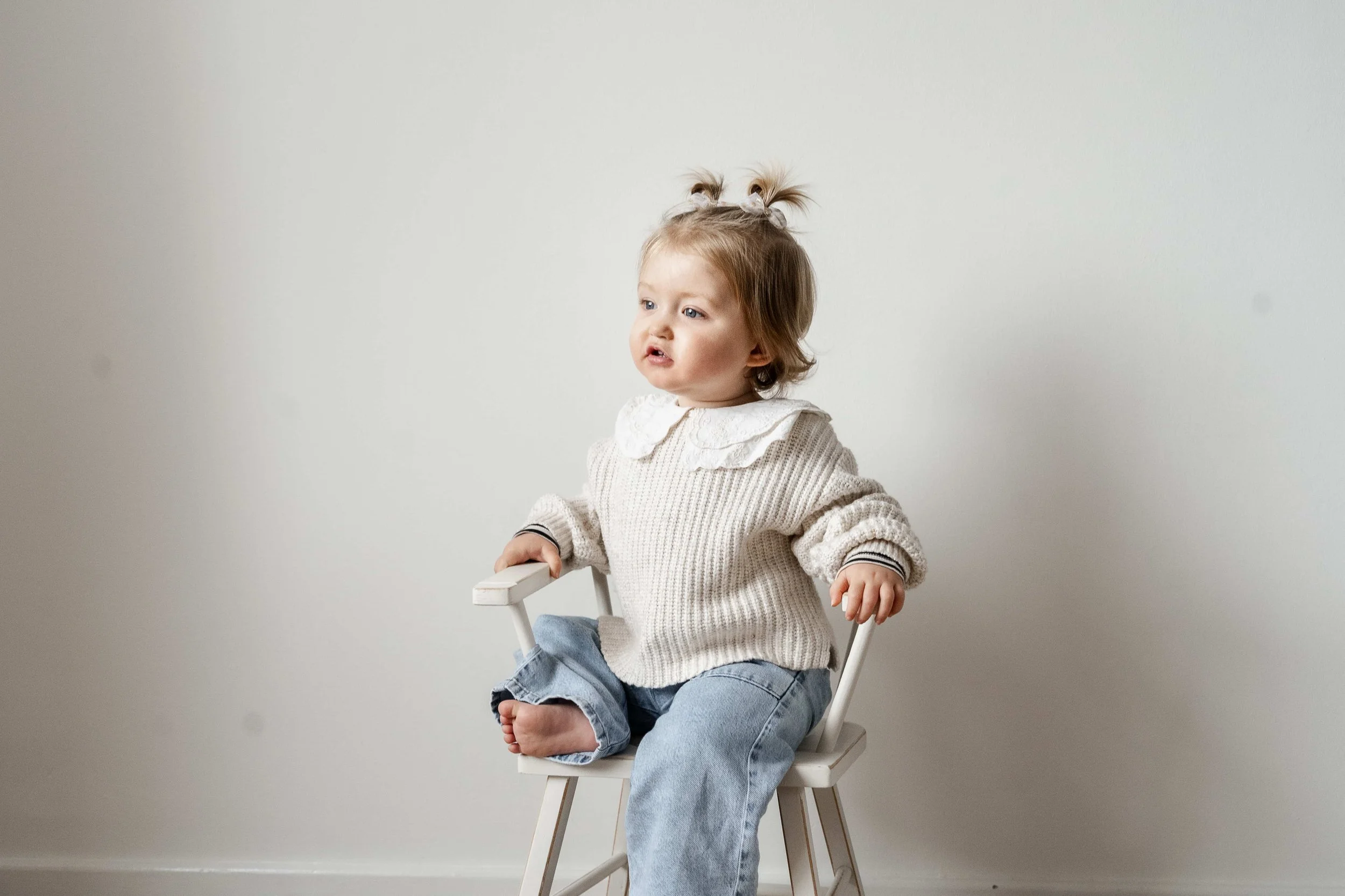 A young girl with light brown hair styled in pigtails with bows, sitting on a white chair against a plain white wall, wearing a knitted sweater and jeans, with her legs crossed and a slightly puzzled expression.