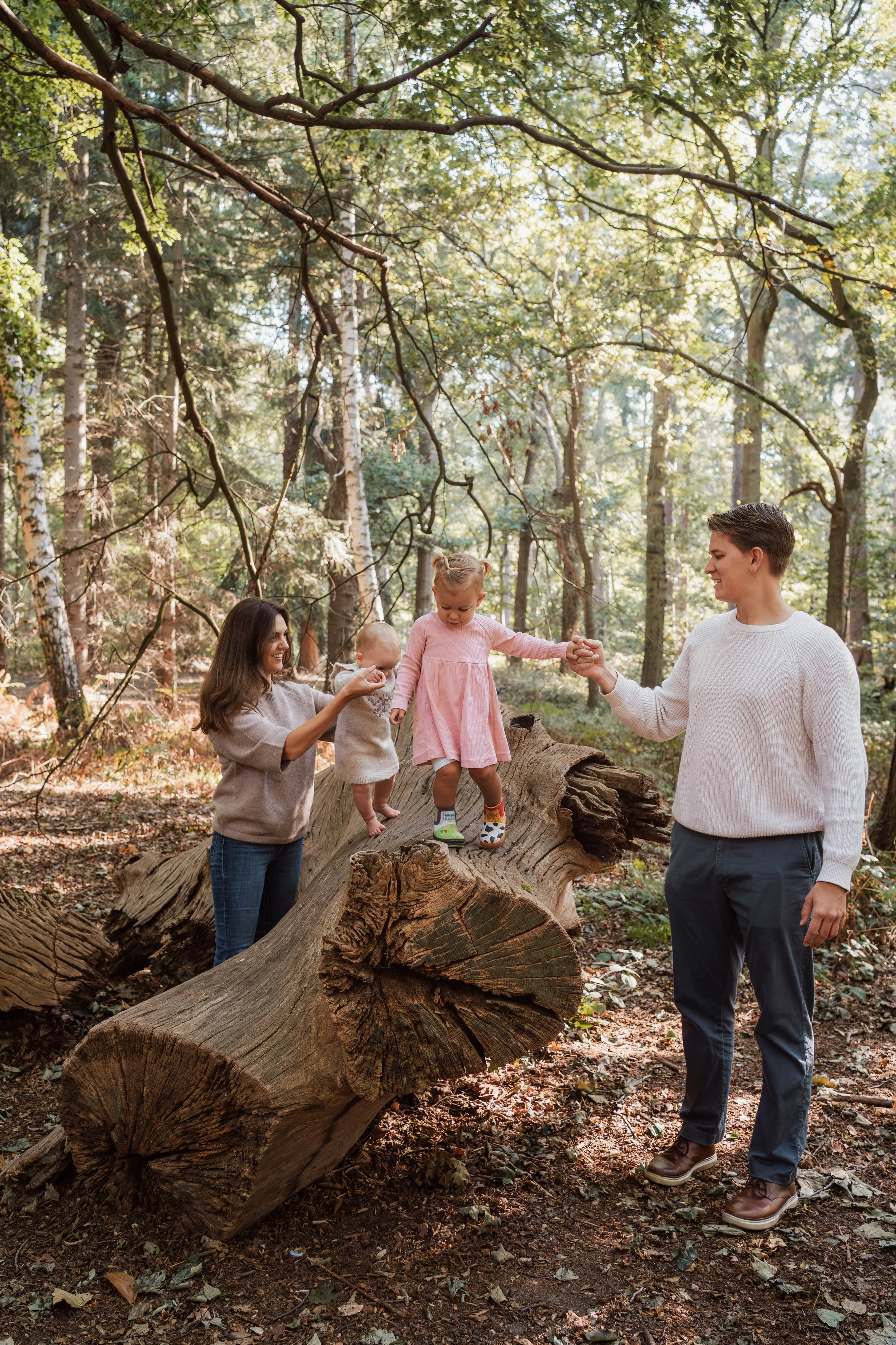 A family of four with two young children outdoors in a forest, with two children standing on a fallen tree, one girl in a pink dress holding a hand of her father, and a woman helping to support a toddler, surrounded by trees and sunlight filtering th