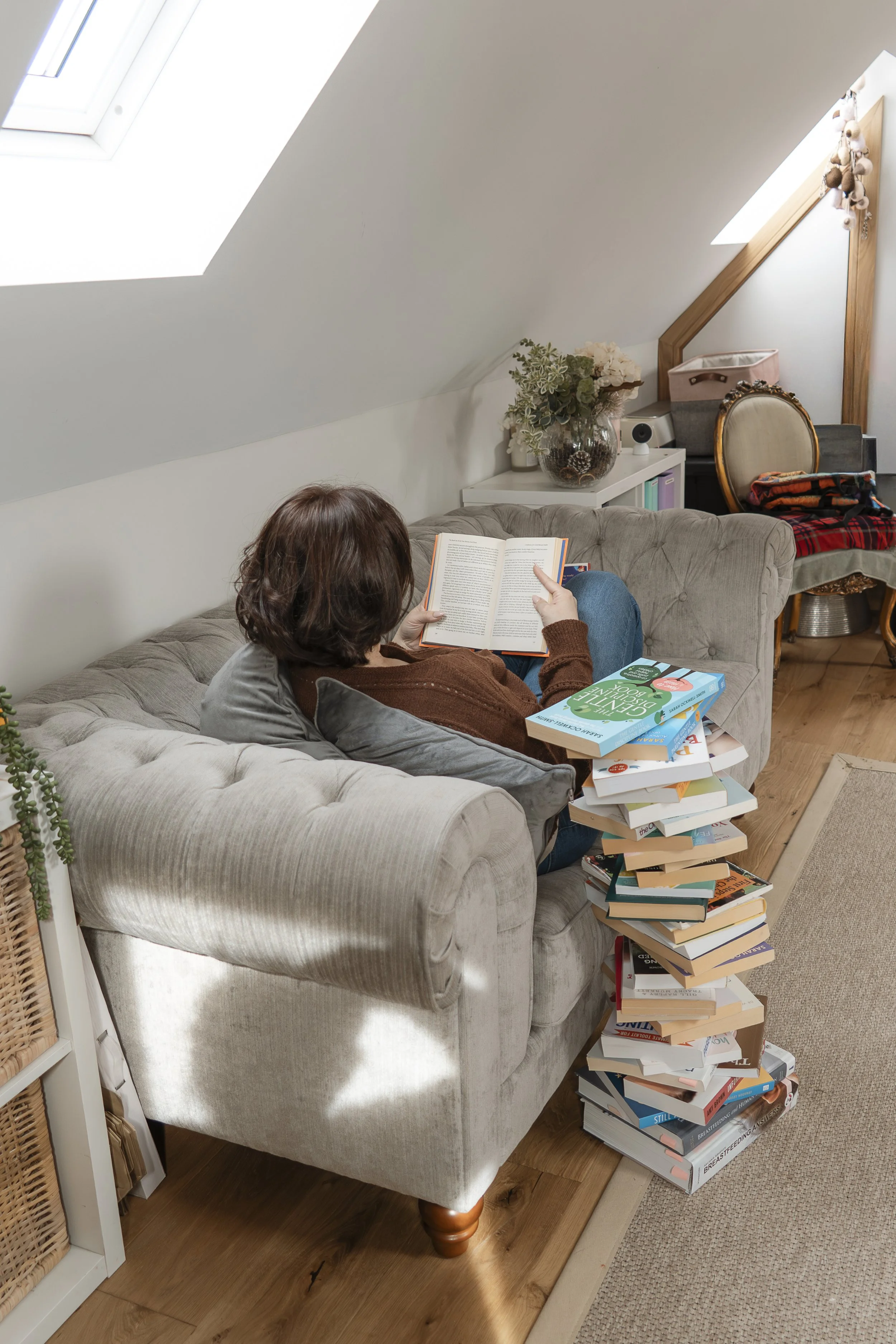 A person with short brown hair sits on a beige tufted sofa, reading a book in a cozy room with sloped ceilings and a skylight, surrounded by a large stack of books.