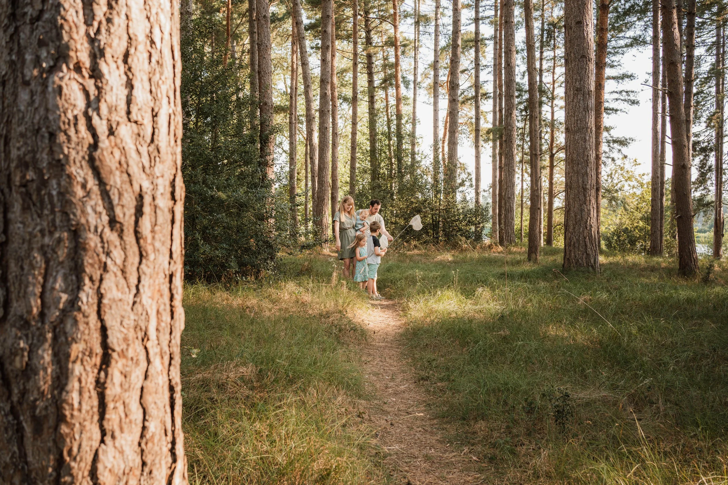 A family of five, including two adults and three children, exploring the woods on a narrow dirt trail during daytime.