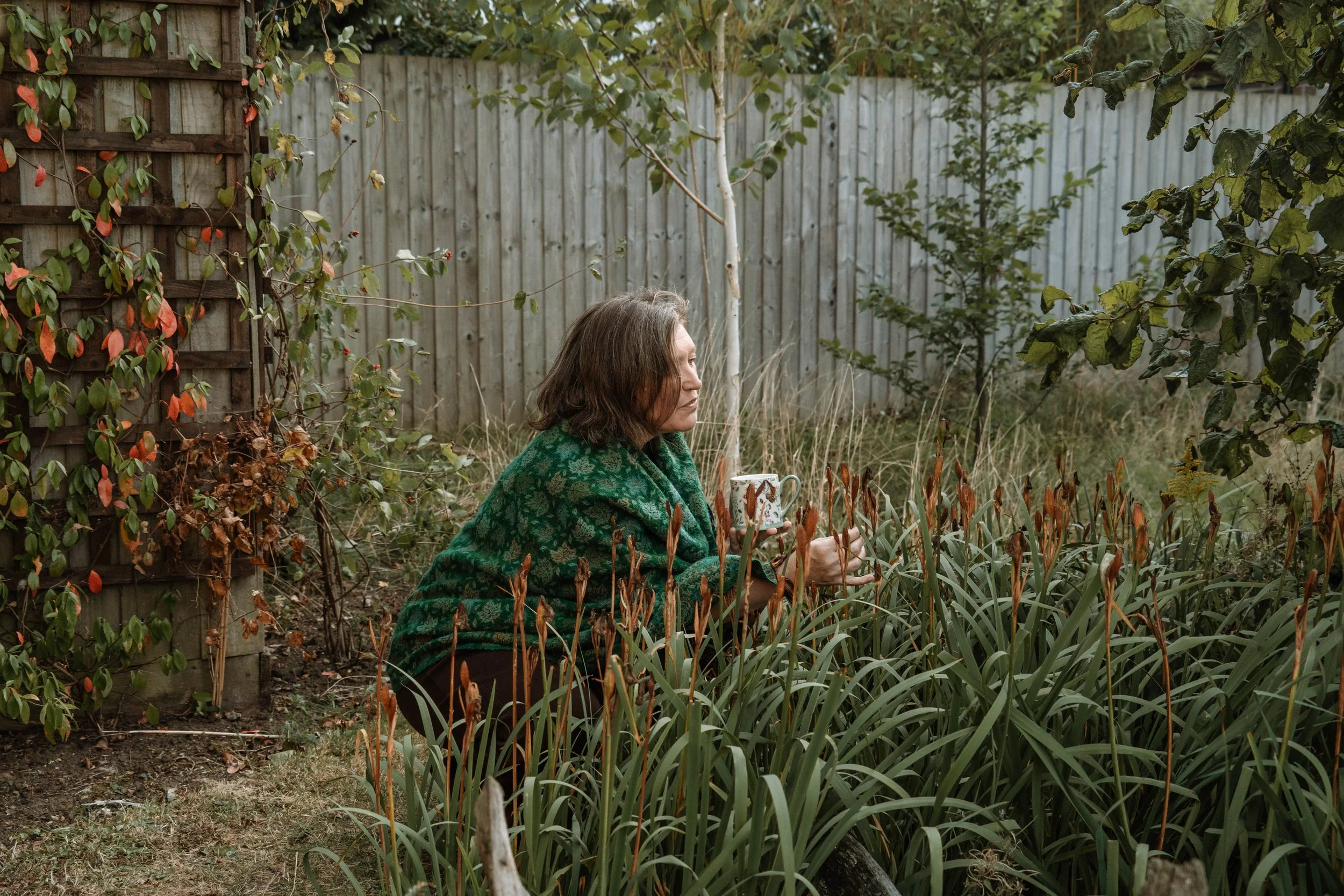 A woman kneeling in a garden with a mug, surrounded by plants and trees, with a wooden fence in the background.
