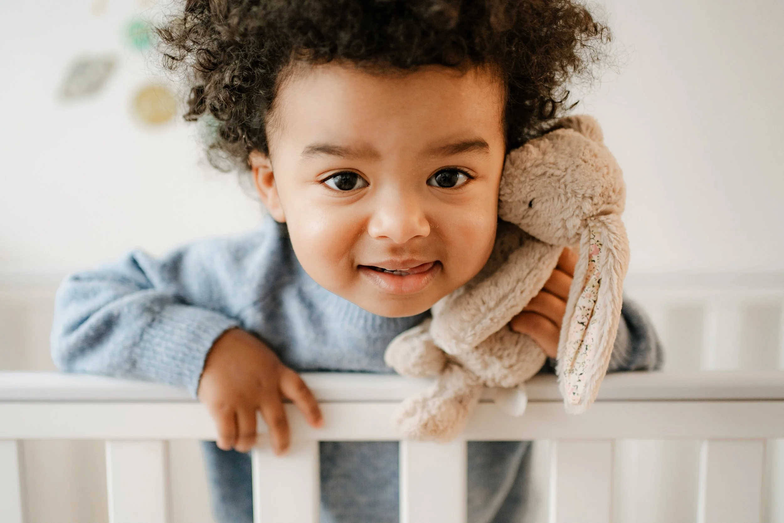 A young child with curly hair leaning over a white crib, holding a plush bunny toy, smiling and looking into the camera in a nursery setting.
