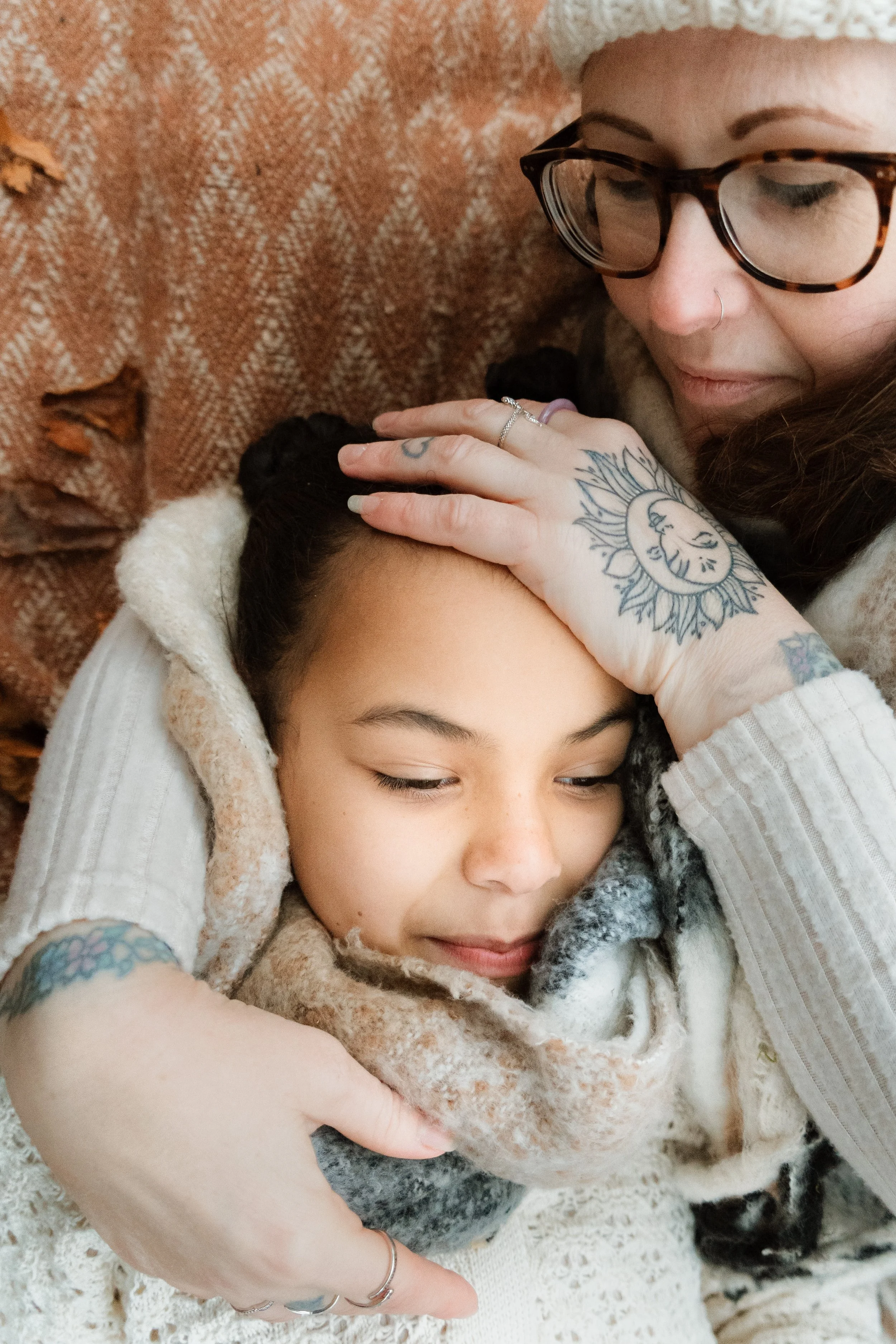 Two women embracing, one lying on a couch with a scarf and the other resting her hand on her head. They appear to be happy and comfortable, surrounded by a warm-toned couch.