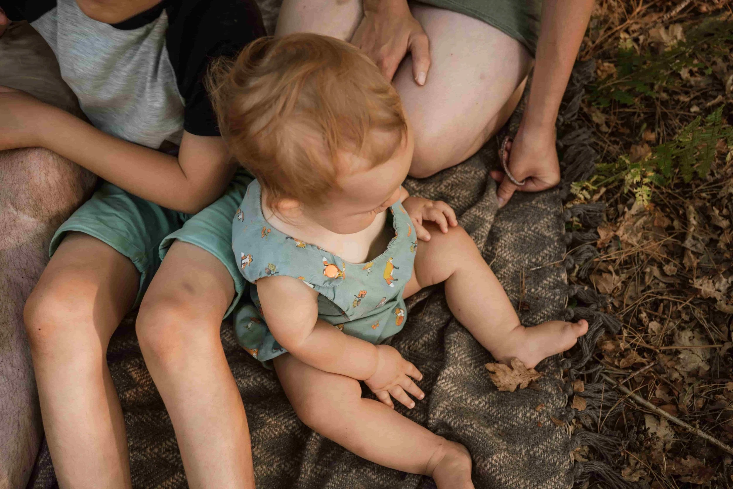 A young child with red hair and light skin, wearing a shirt with foxes, sitting on a blanket outdoors surrounded by fallen leaves. A woman with light skin and wearing a green shirt and shorts is lying beside the child, with her head resting on her ar