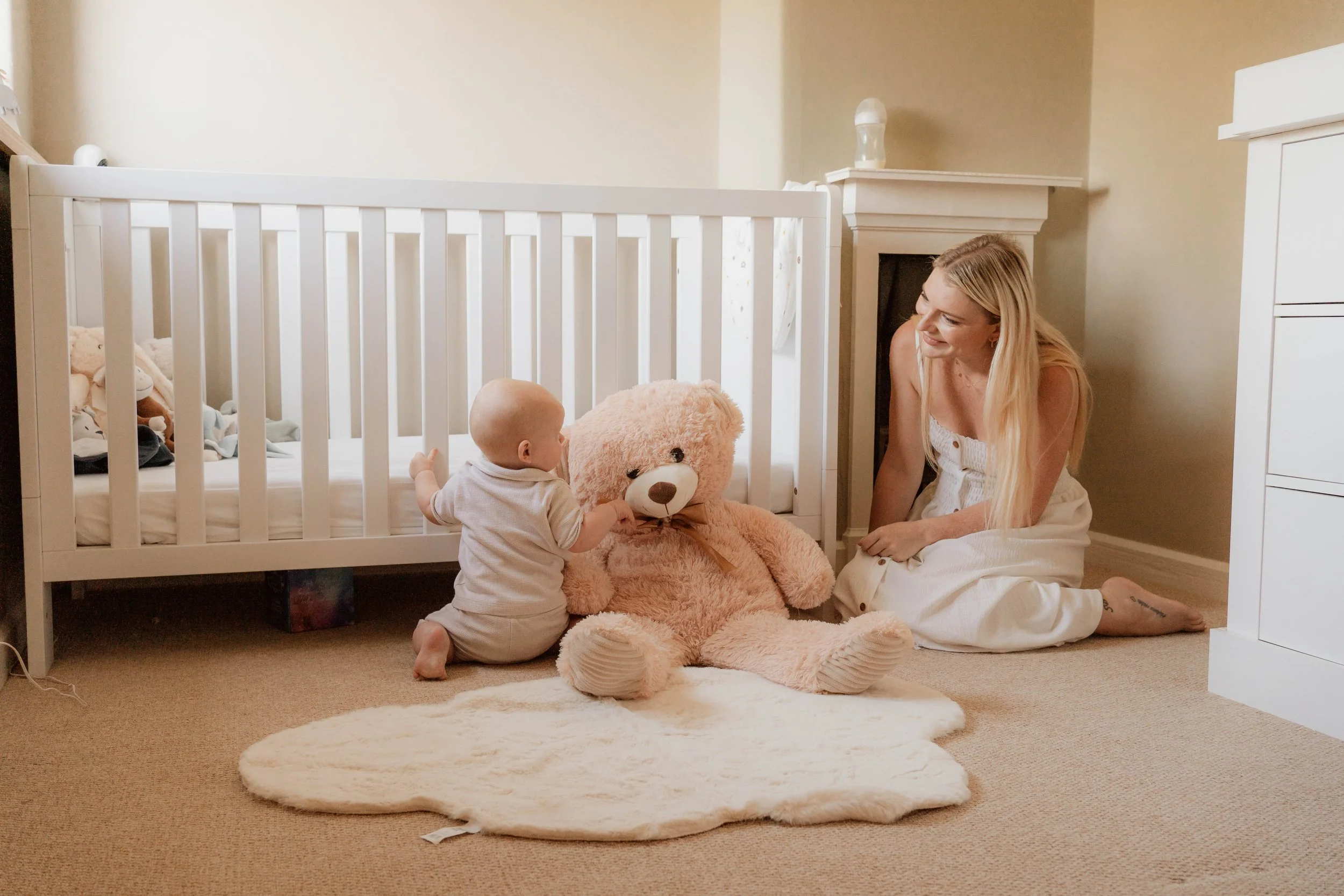 A woman and a baby sitting on the floor near a bed and a giant teddy bear in a cozy nursery.