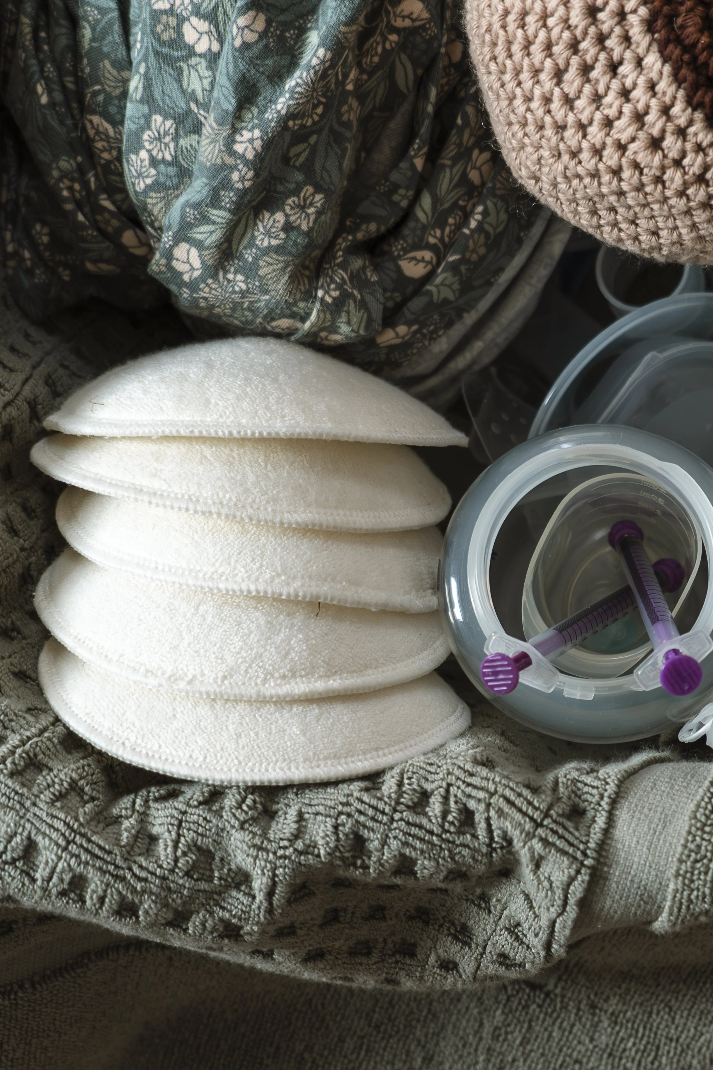 Disposable breast shields stacked next to a plastic container with purple-capped syringes on a textured gray cloth, with clothing and a pink knit hat in the background.