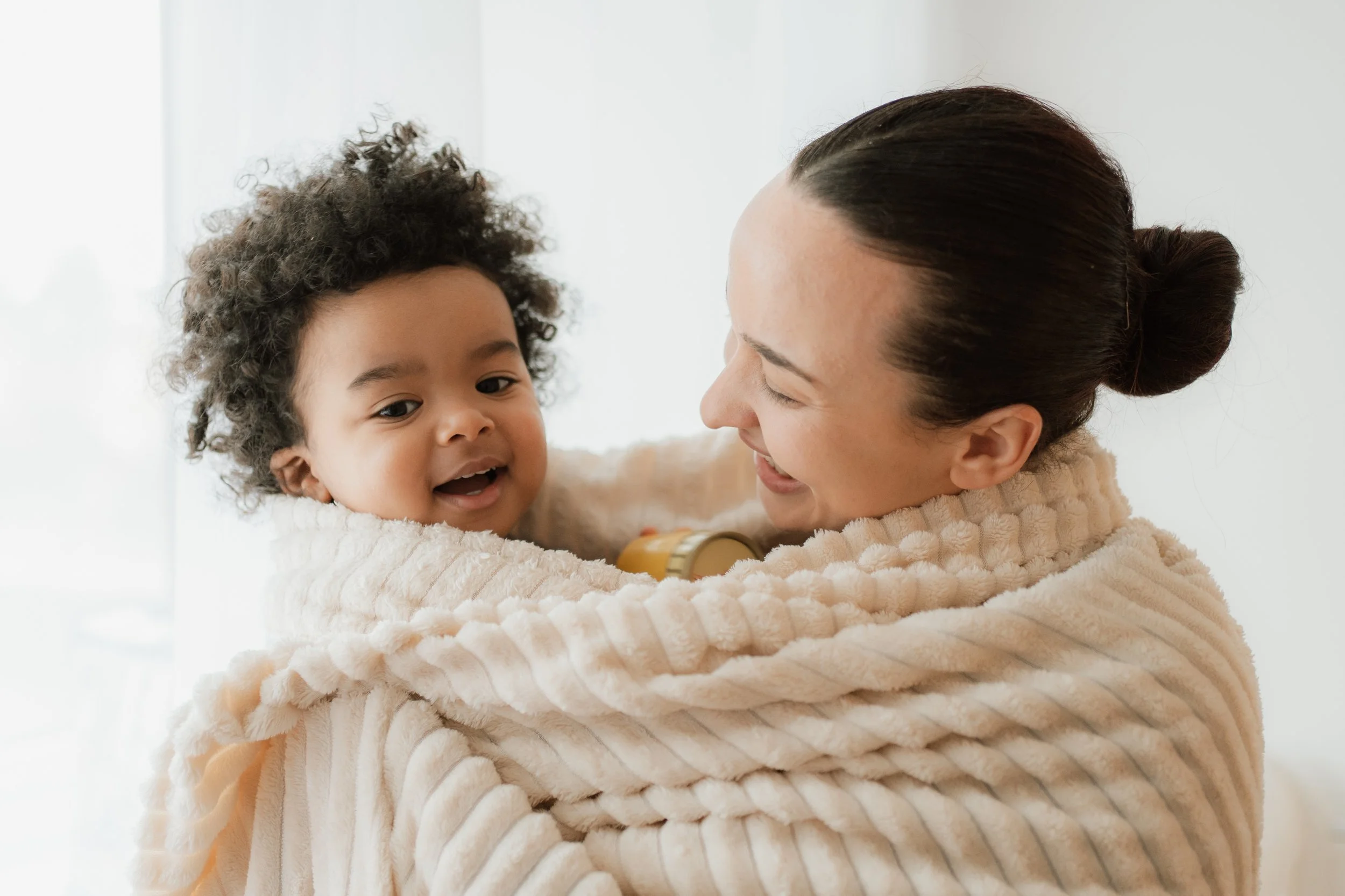 A woman holding a smiling young child in a cozy beige blanket, with a bright background.