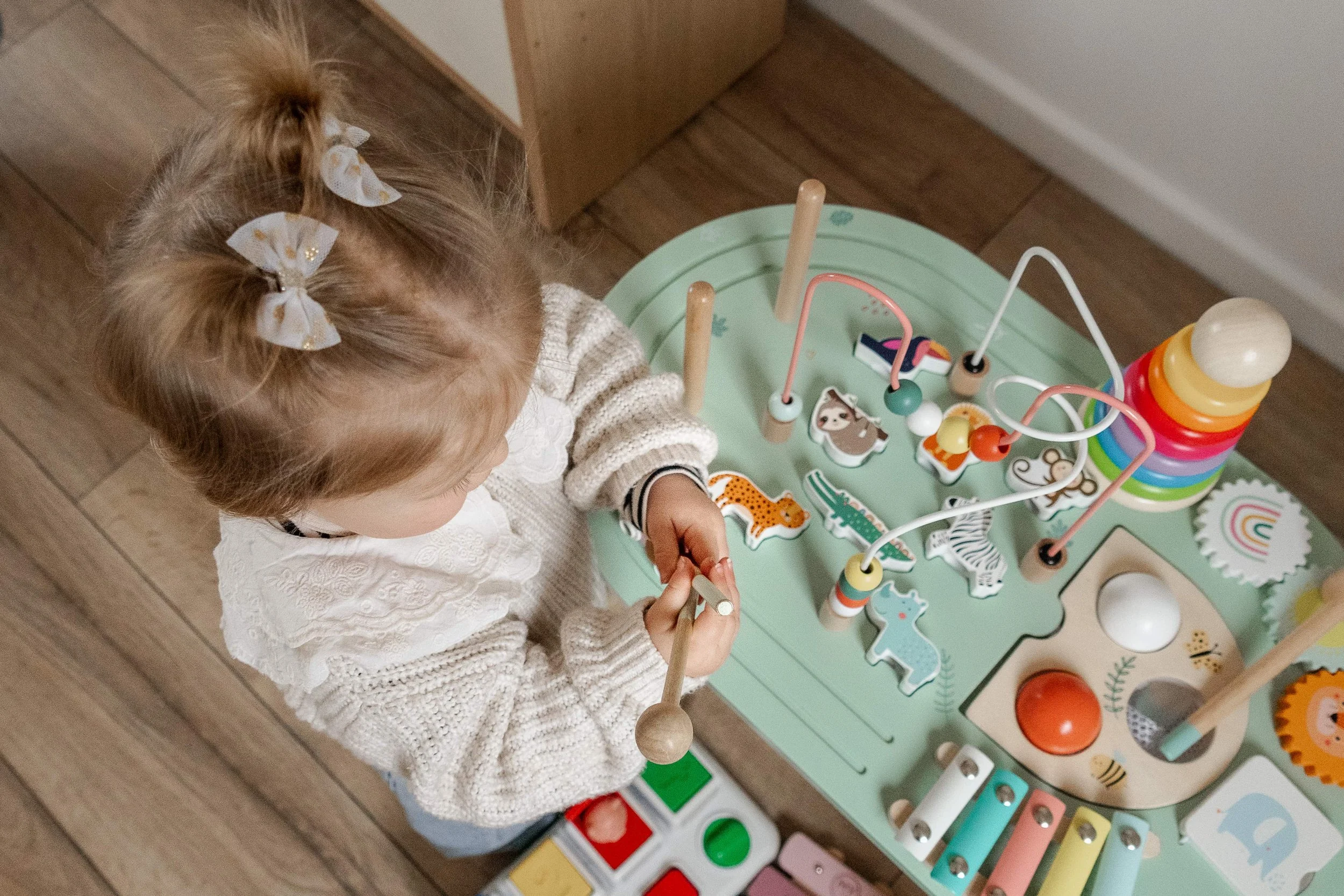 A young girl with blonde hair tied in pigtails with white bows, wearing a beige sweater, is standing at a play table with colorful toys and activities.
