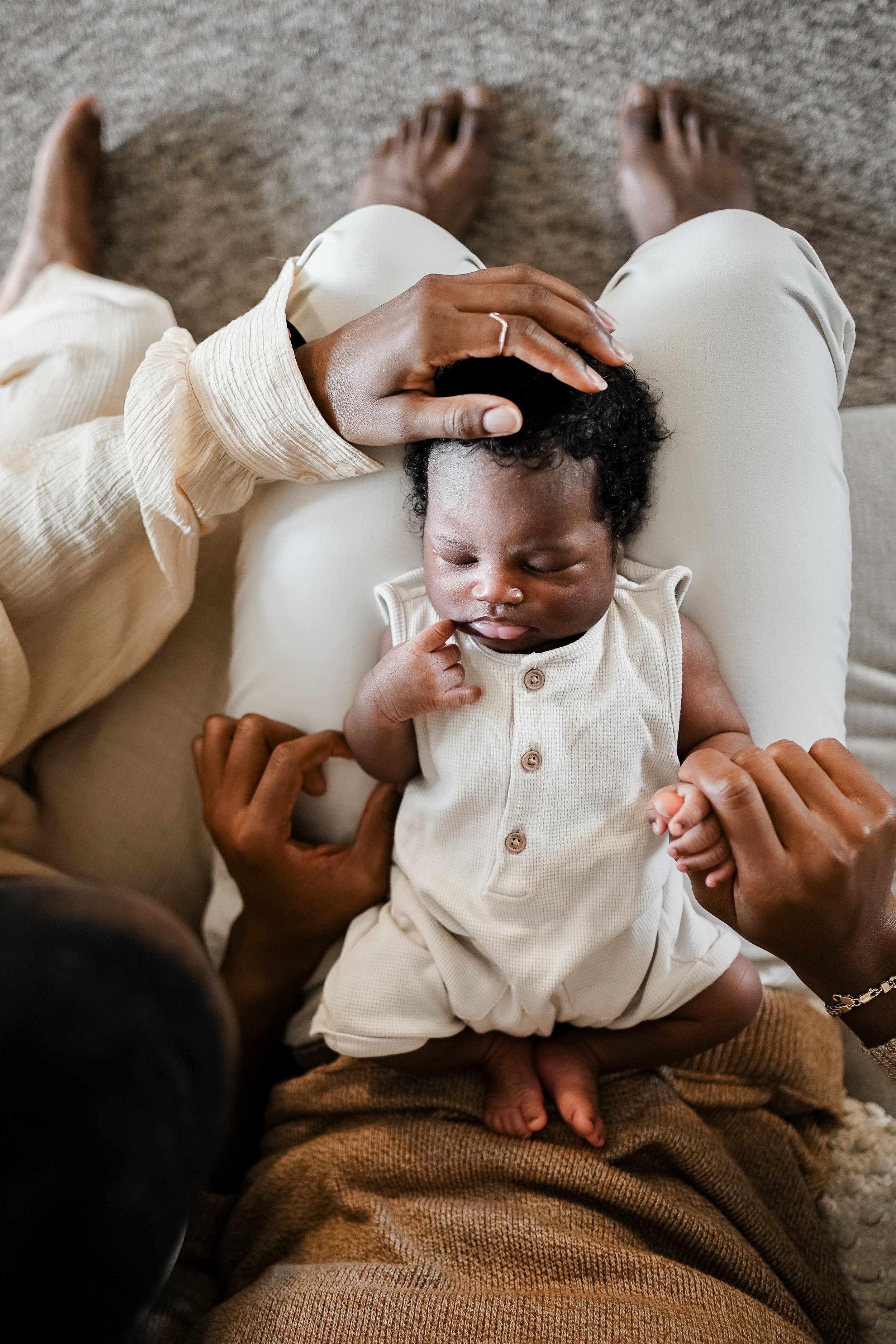A person sitting on the floor, holding a sleeping baby in their lap. The baby is wearing a cream-colored outfit and is resting peacefully with one hand near their mouth. The person is gently touching the baby's head with one hand and holding the baby